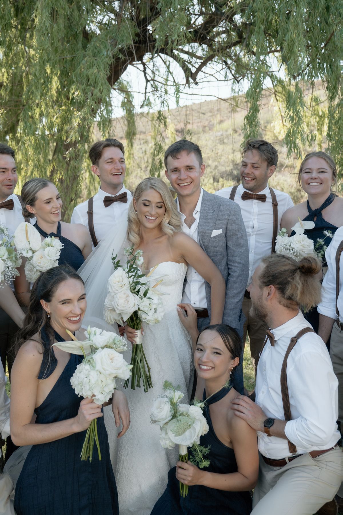 Smiling bride and groom surrounded by wedding party in navy and grey attire under a willow tree.
