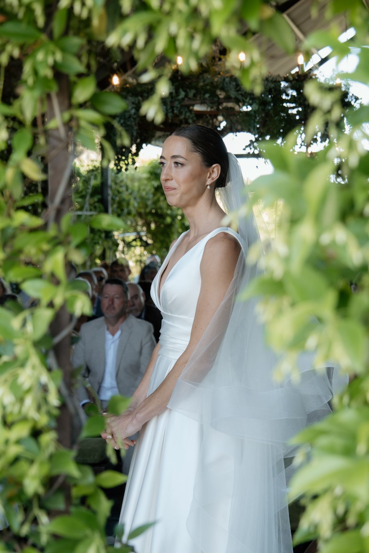 Beautiful bride in white gown and veil stands framed by lush green foliage during outdoor ceremony.