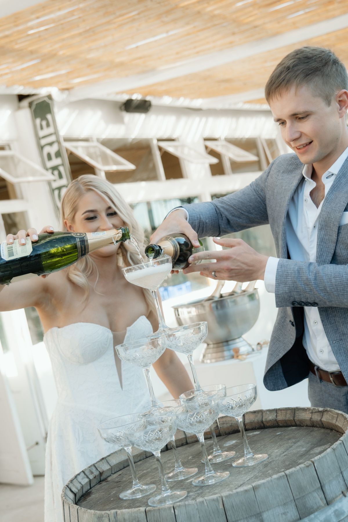 Smiling bride and groom pour champagne tower together at elegant outdoor wedding reception.