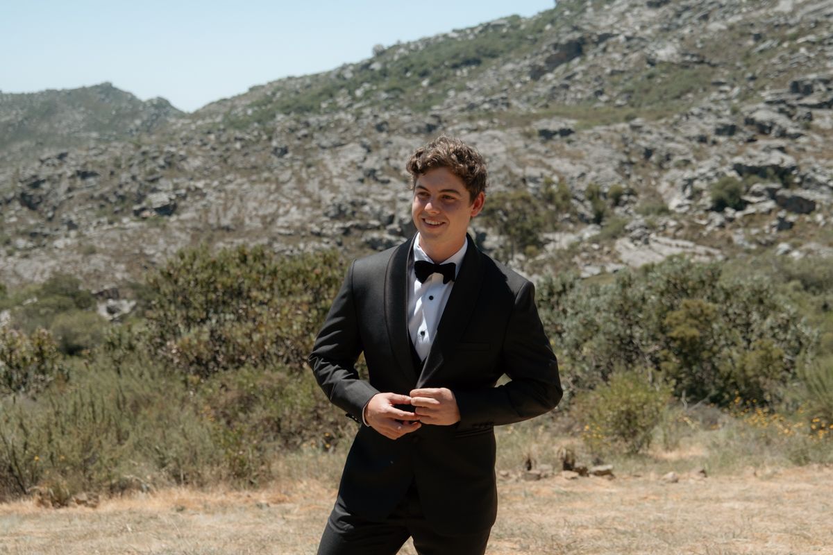Young man in black tuxedo and bow tie standing outdoors with rocky hills and shrubs in the background.