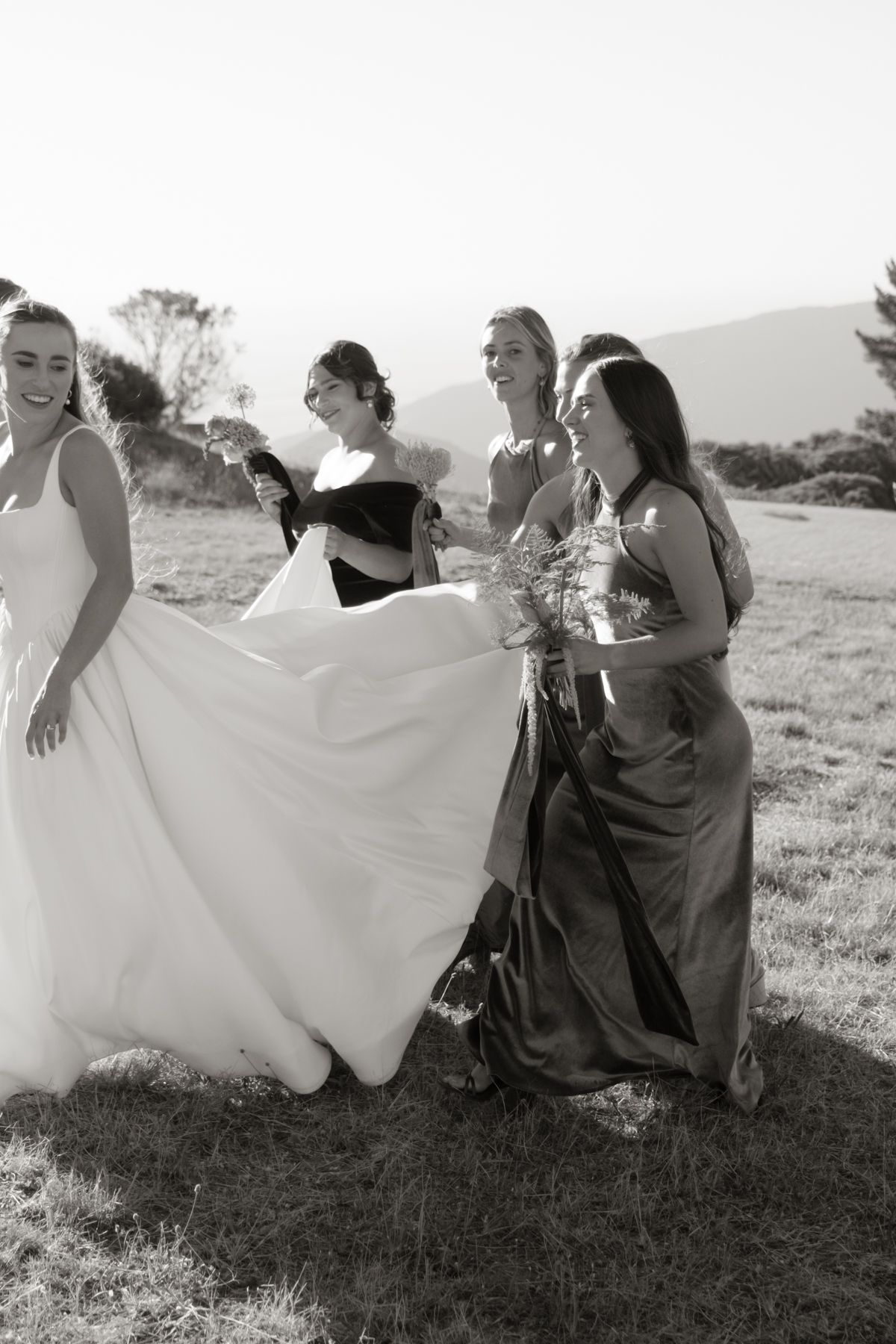 Bridesmaids in olive and grey gowns help bride with her white dress train outdoors in black and white photo.