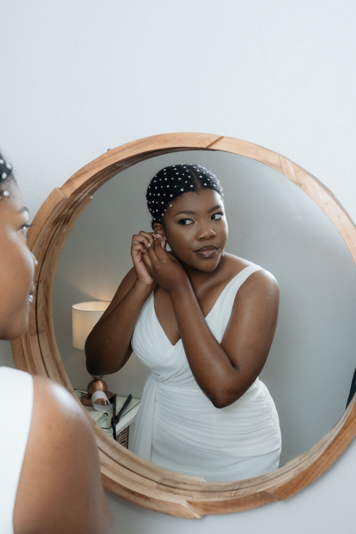 A Black woman in a white dress adjusts her earring while looking into a round wooden-framed mirror.