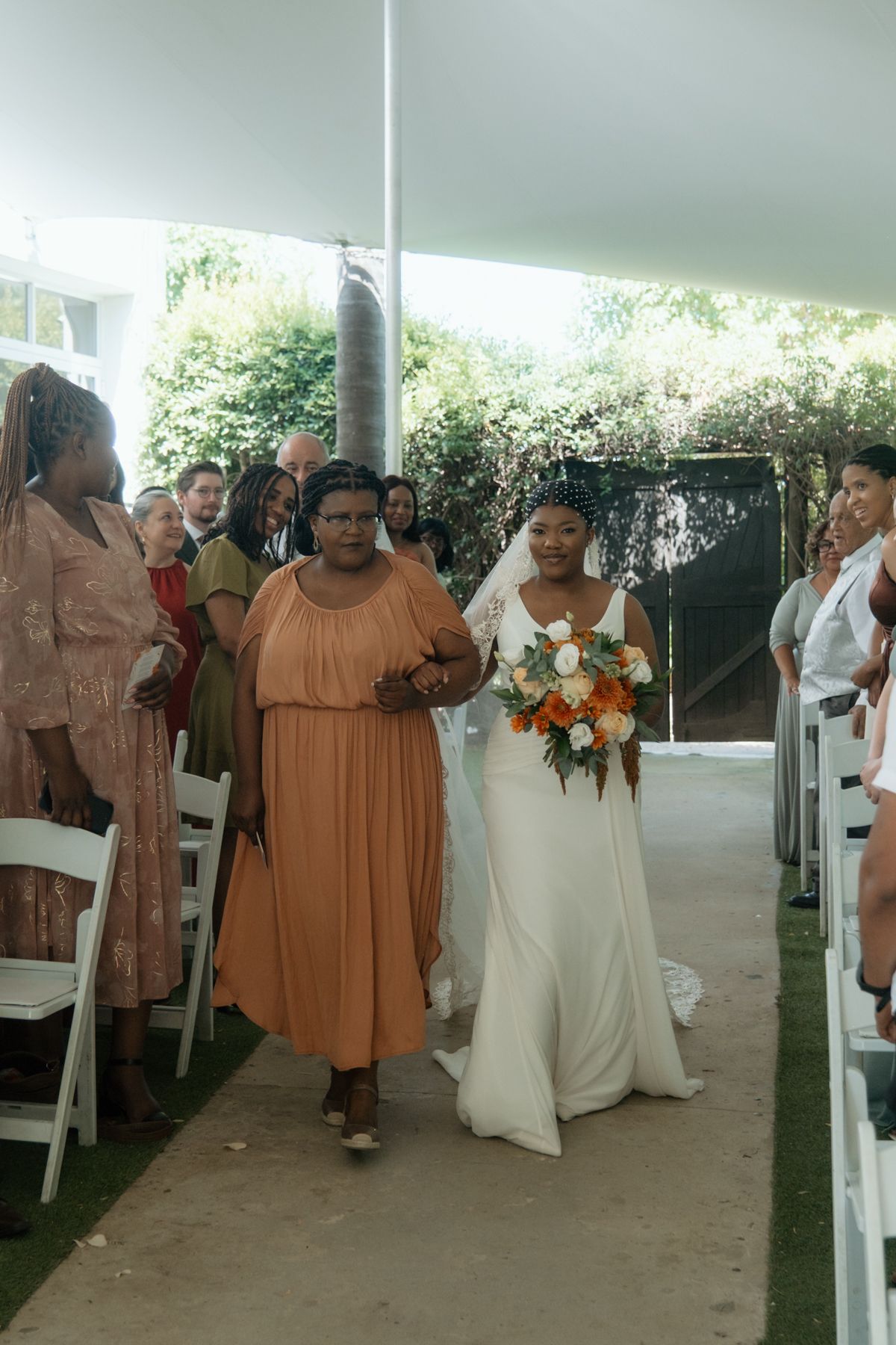 Bride in white gown walks down the aisle with escort at outdoor wedding ceremony under white tent.
