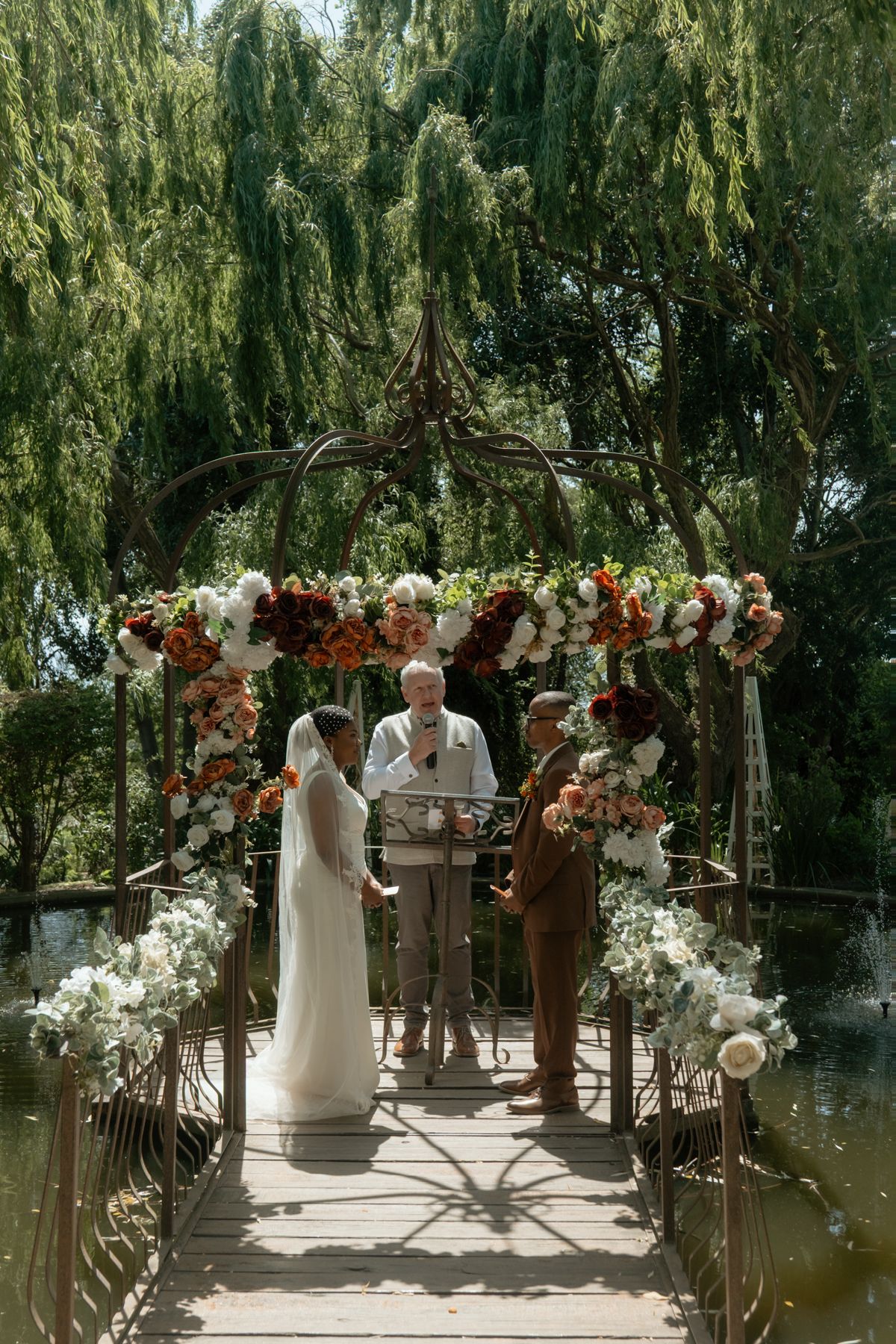 Bride and groom exchange vows at floral arch gazebo over pond with officiant.