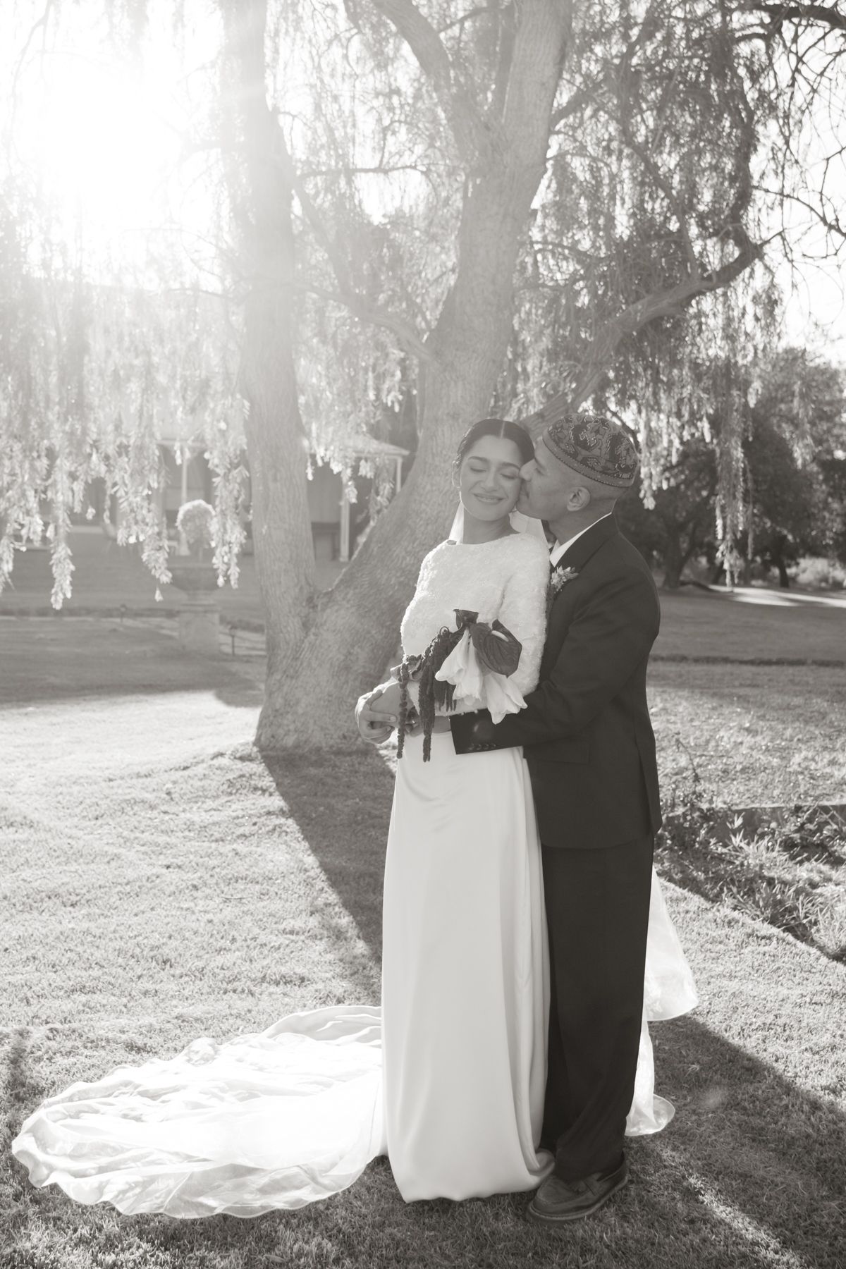Black and white photo of bride and groom embracing under a weeping willow tree at wedding.