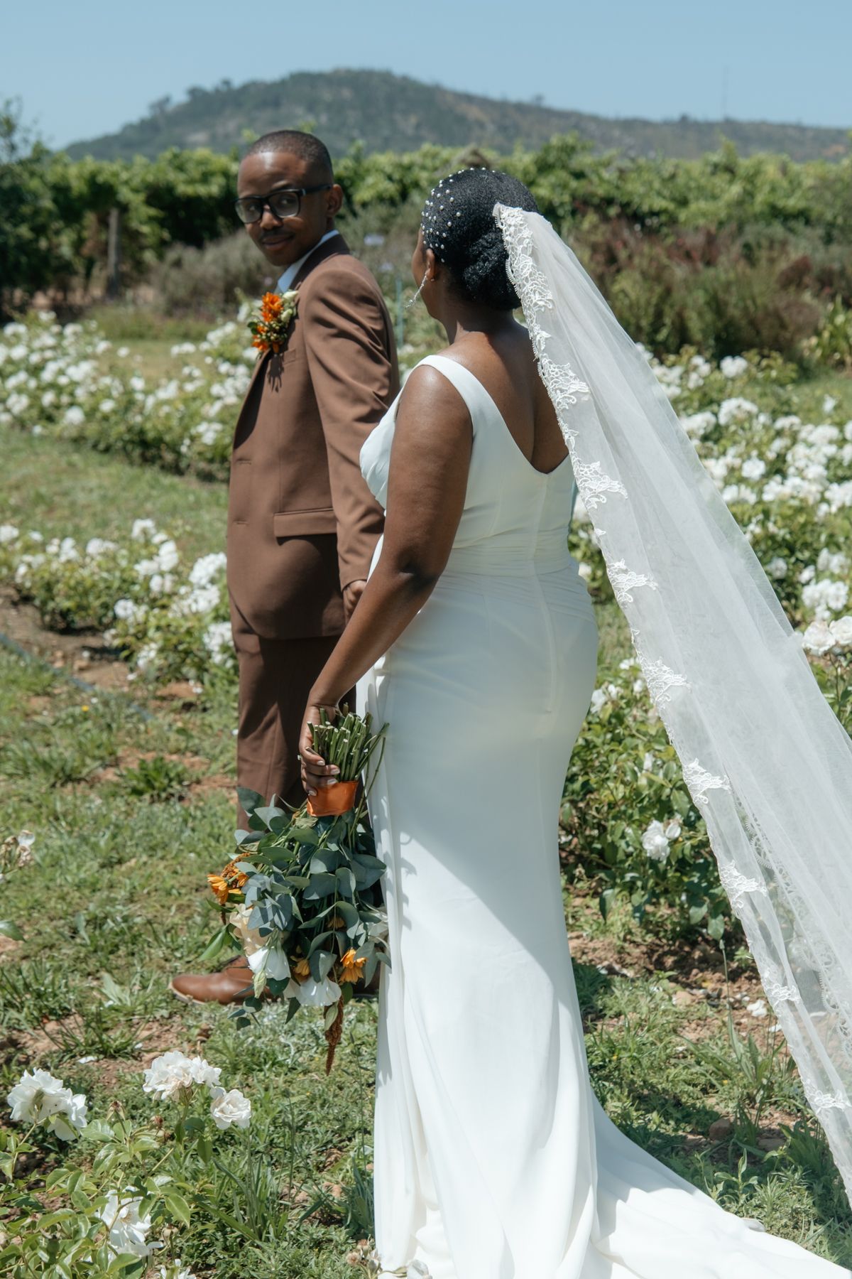 Bride in white gown with lace veil and groom in brown suit posing in a rose garden with mountain views.