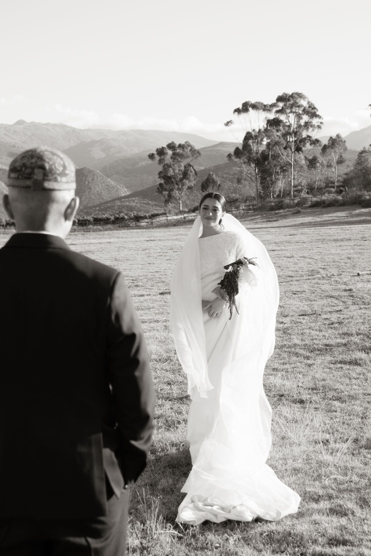 Black and white first look photo of bride in veil holding bouquet facing groom with mountain backdrop.
