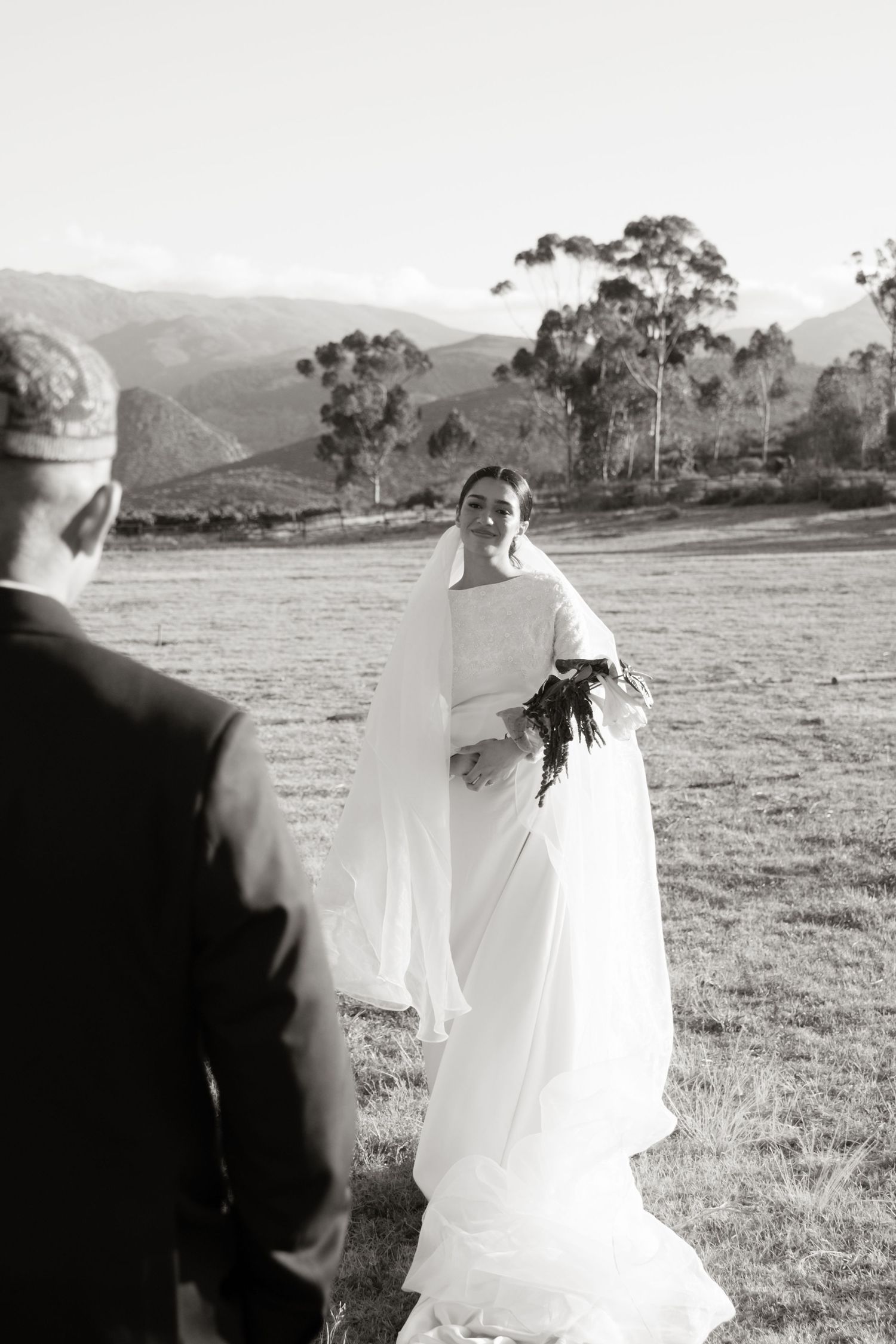 Black and white photo of smiling bride in flowing white gown holding bouquet facing groom in mountain landscape.