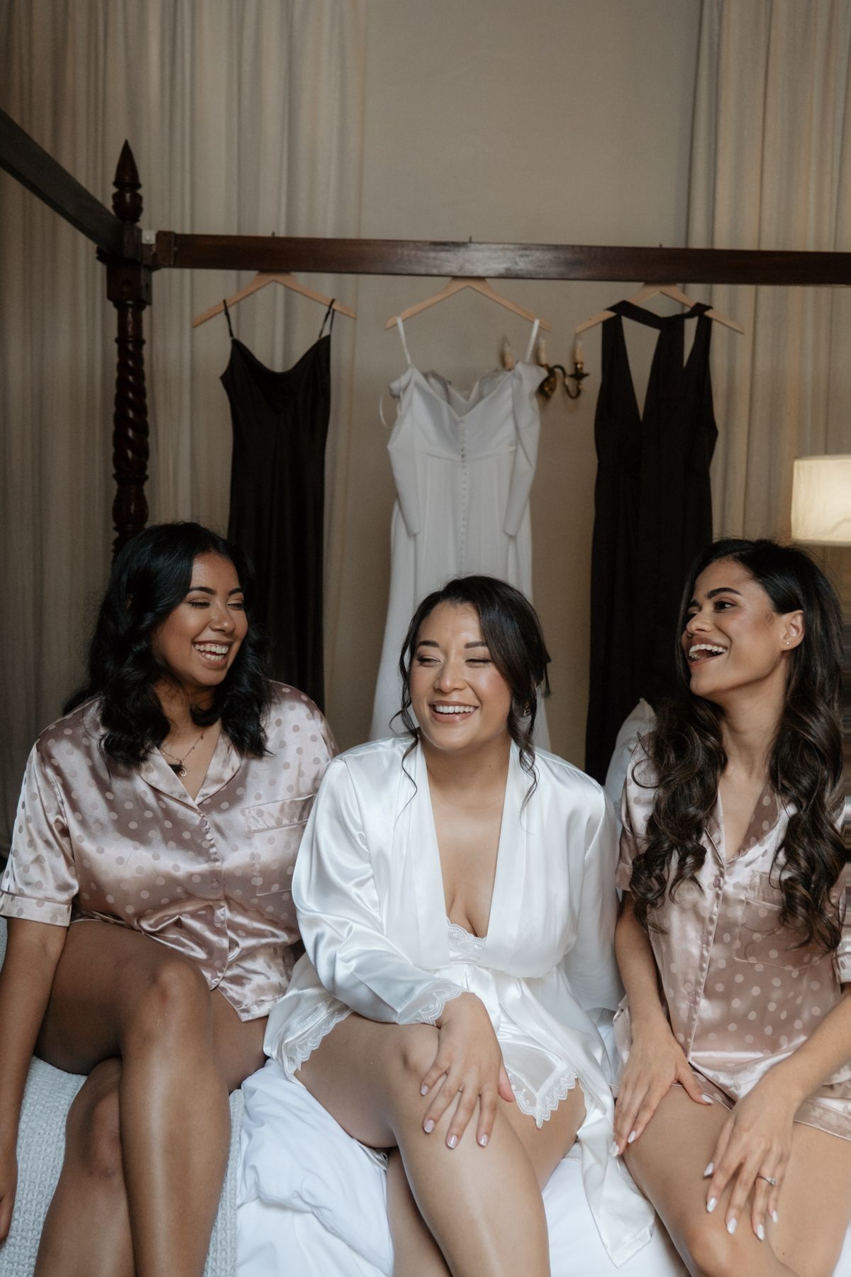 Bride in white robe with two bridesmaids in satin pajamas laughing during wedding preparations.