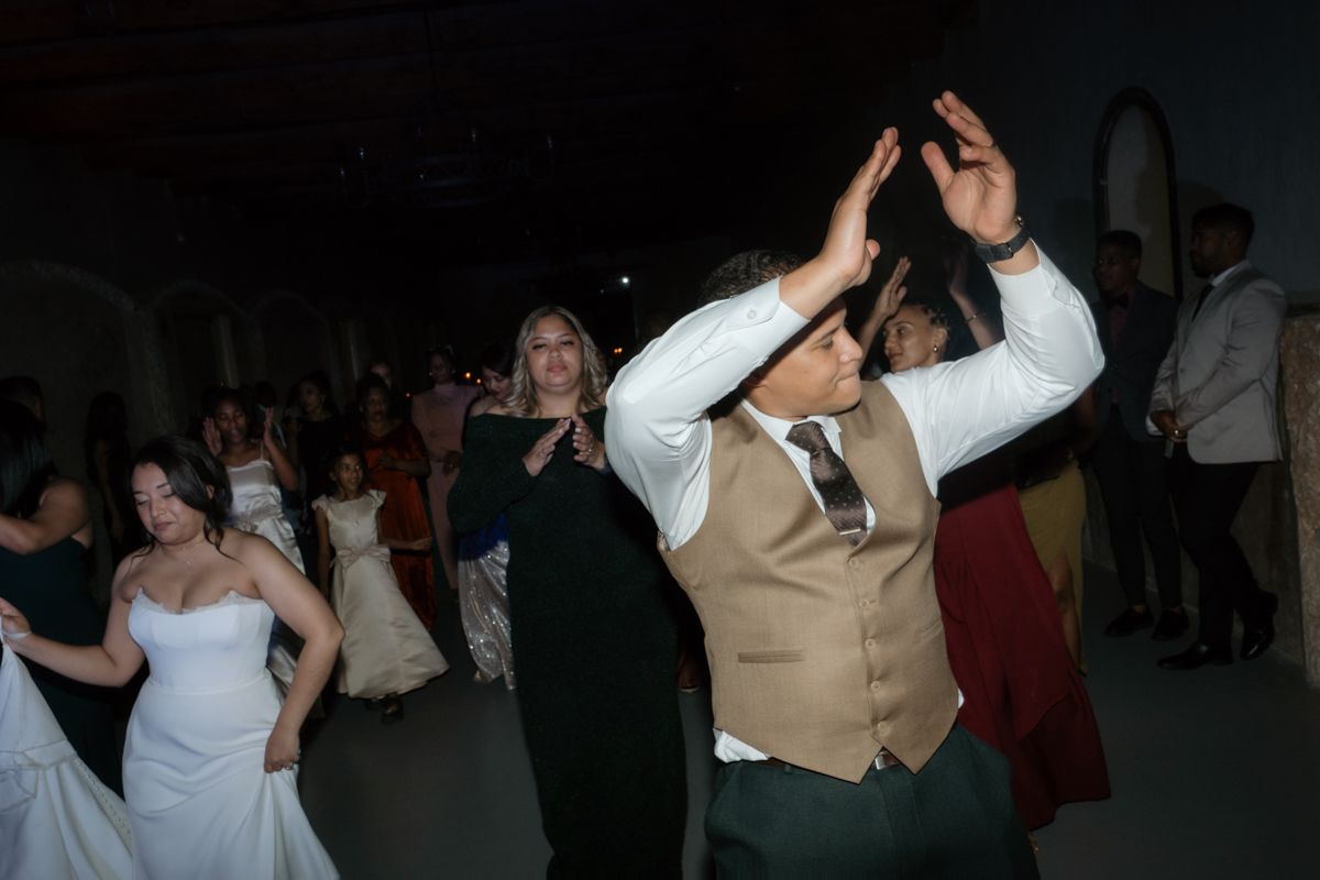 Energetic wedding guests including groom in tan vest dancing with raised arms at lively reception.