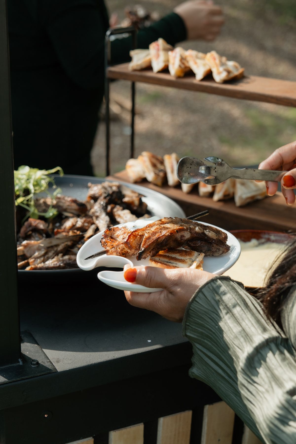 Person serving grilled meat onto a white plate at an outdoor wedding buffet.
