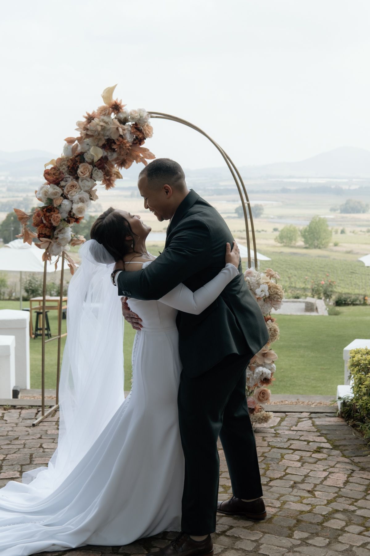 Bride and groom share first kiss under floral arch at outdoor wedding ceremony with scenic vineyard views.