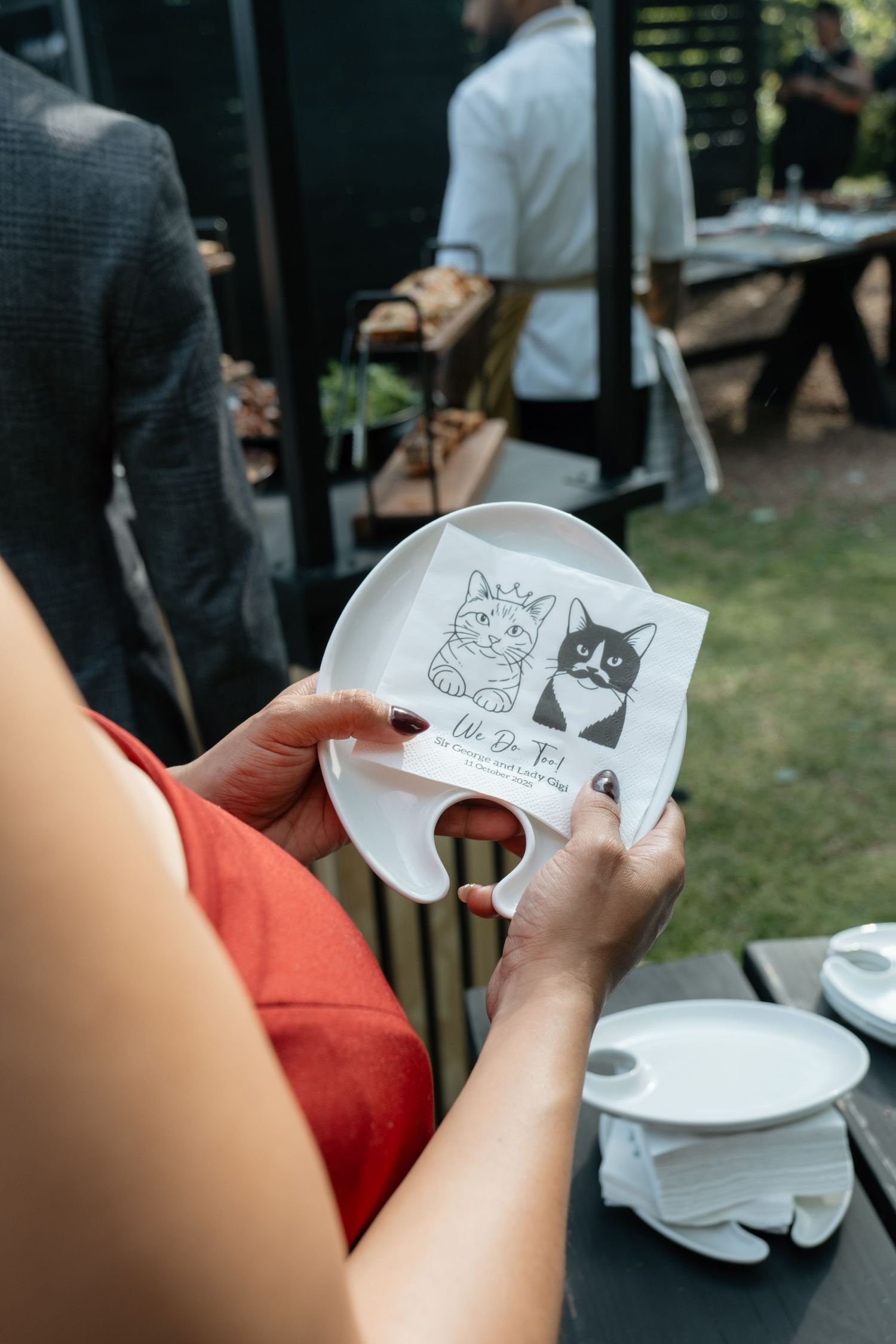 Wedding guest holds white plate with cat-illustrated napkin at outdoor garden reception.