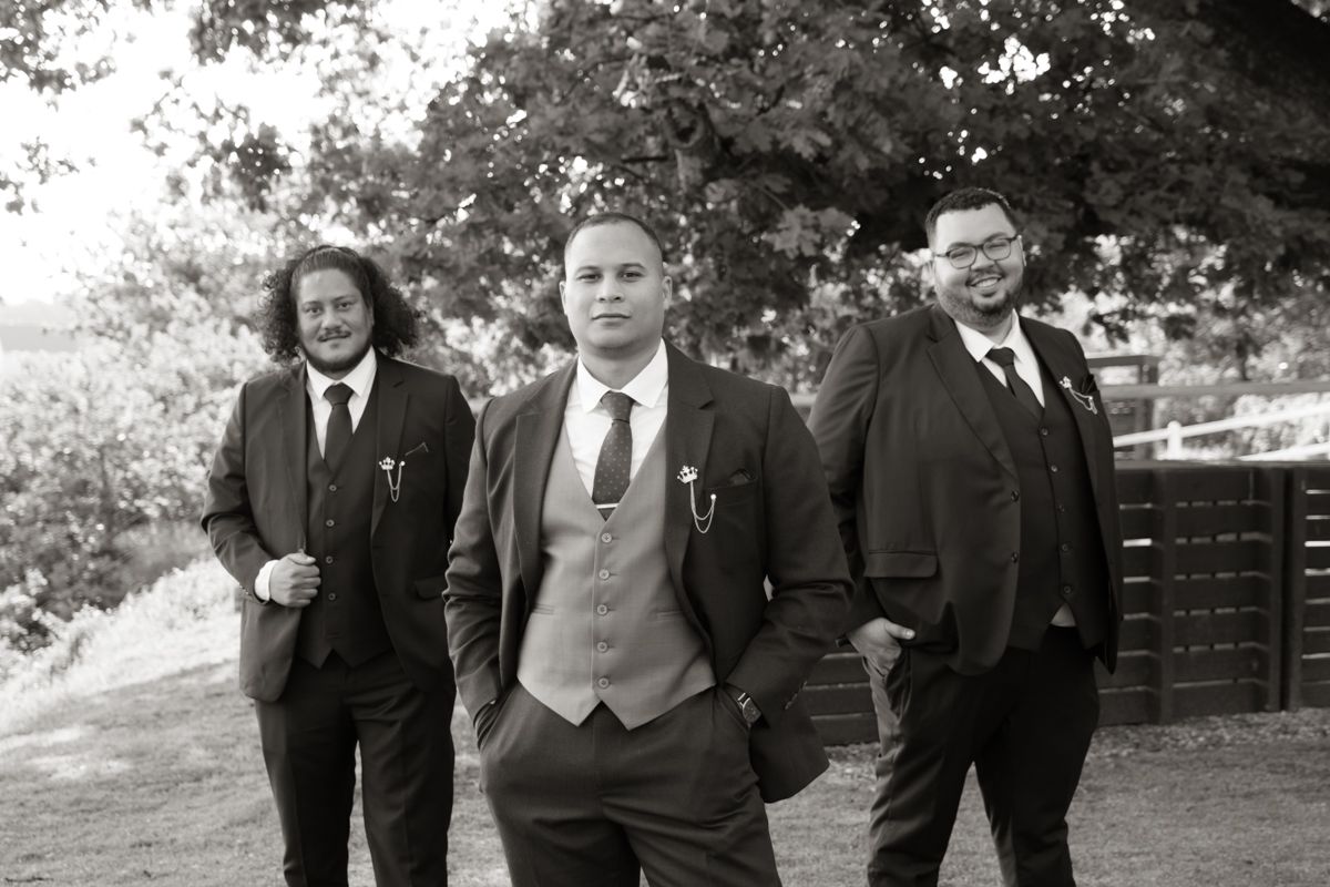 Three groomsmen in dark suits and ties pose outdoors under trees in classic black and white wedding photo.