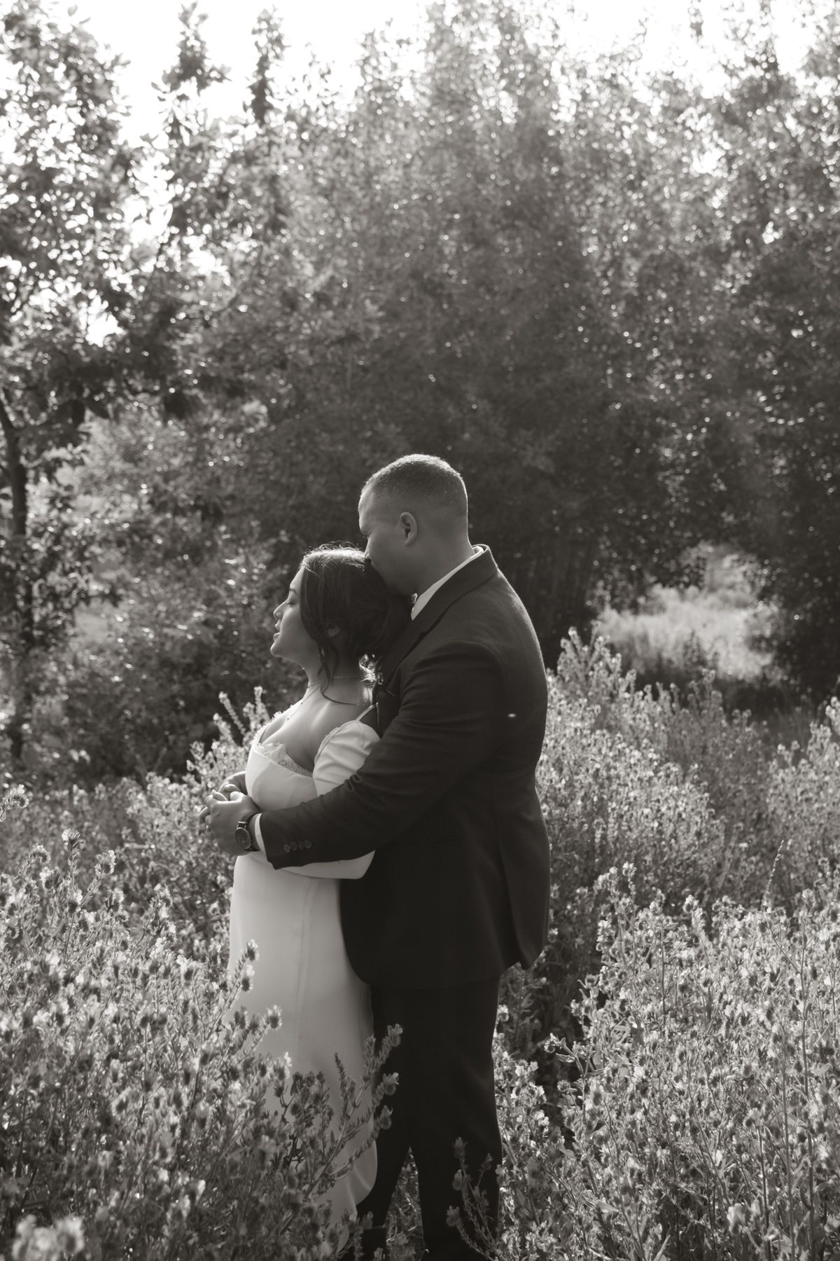 Black and white photo of a bride and groom embracing in a lush garden setting on their wedding day.