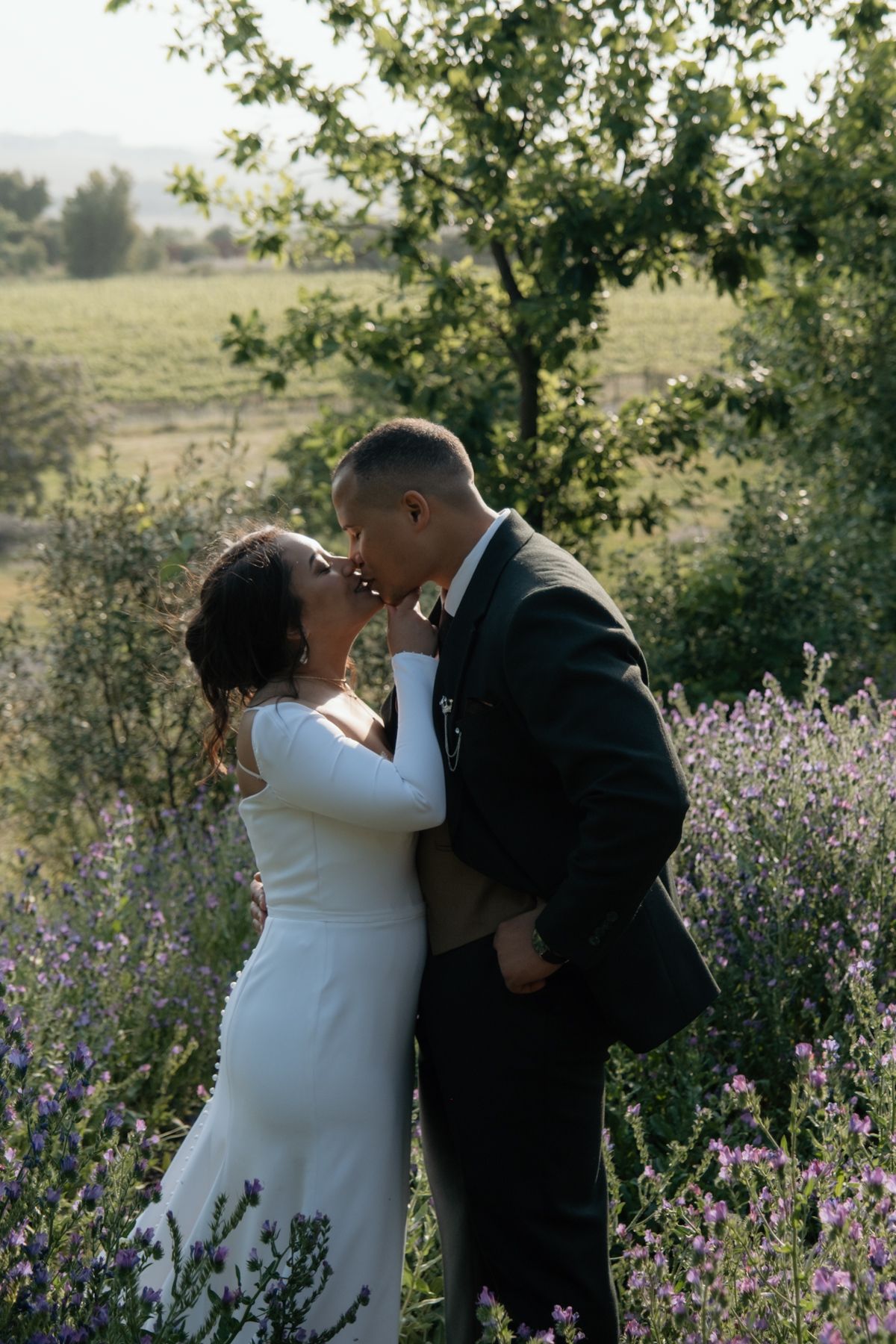 Bride and groom sharing a romantic kiss surrounded by blooming lavender in a scenic outdoor landscape.