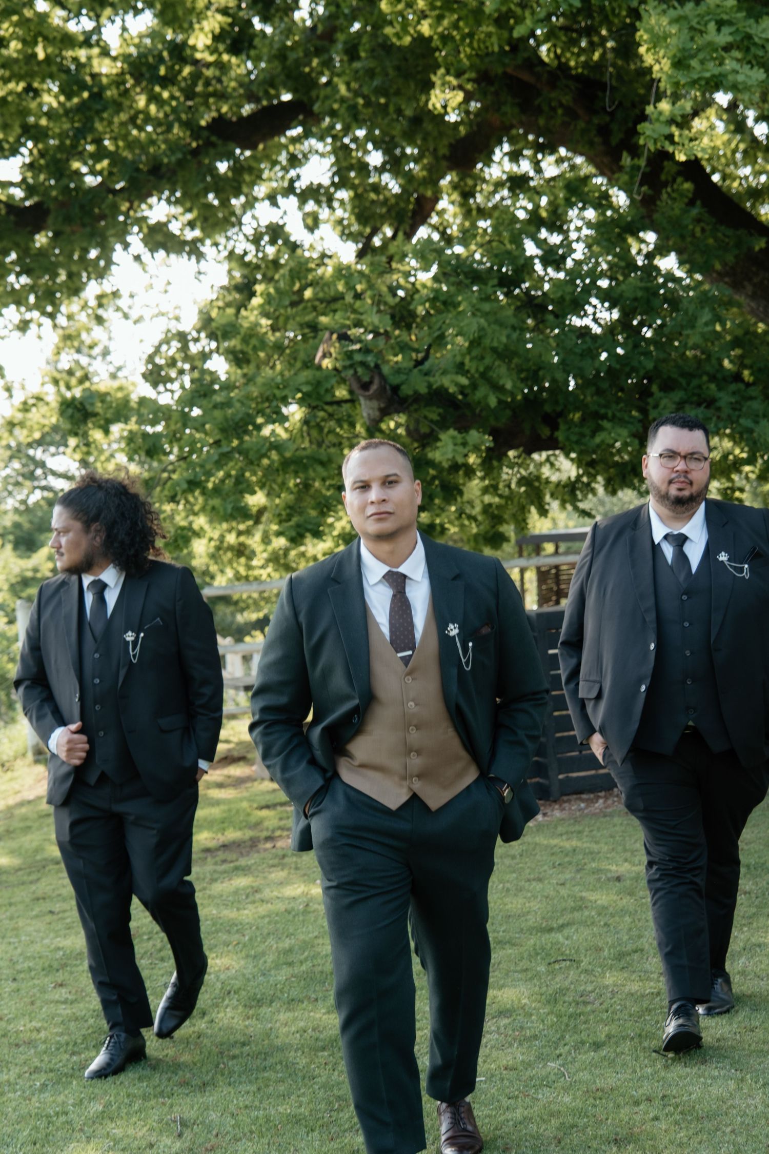 Groom and groomsmen in dark suits walk confidently across green lawn under large tree.