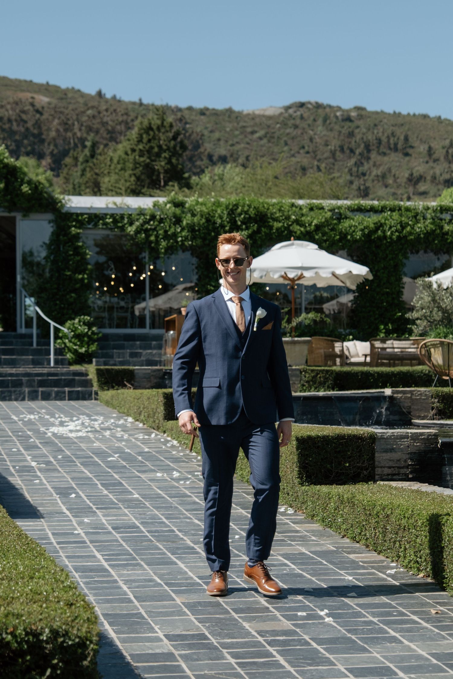 Dapper groom in navy blue suit and tan shoes walks confidently along garden pathway on sunny wedding day.