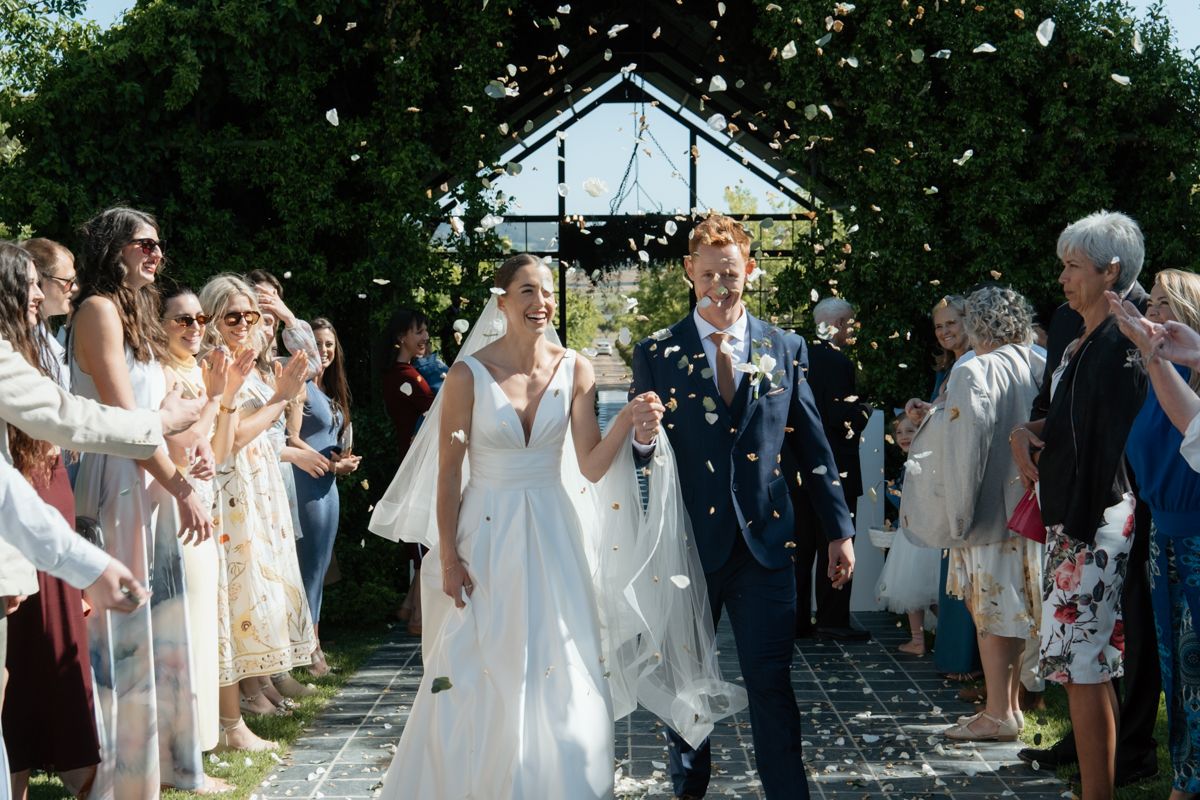 Bride and groom walking through confetti shower after outdoor wedding ceremony with cheering guests.