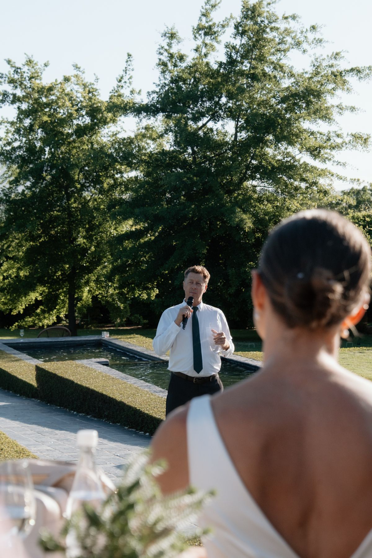 Man delivers outdoor wedding speech with microphone while bride listens in foreground.
