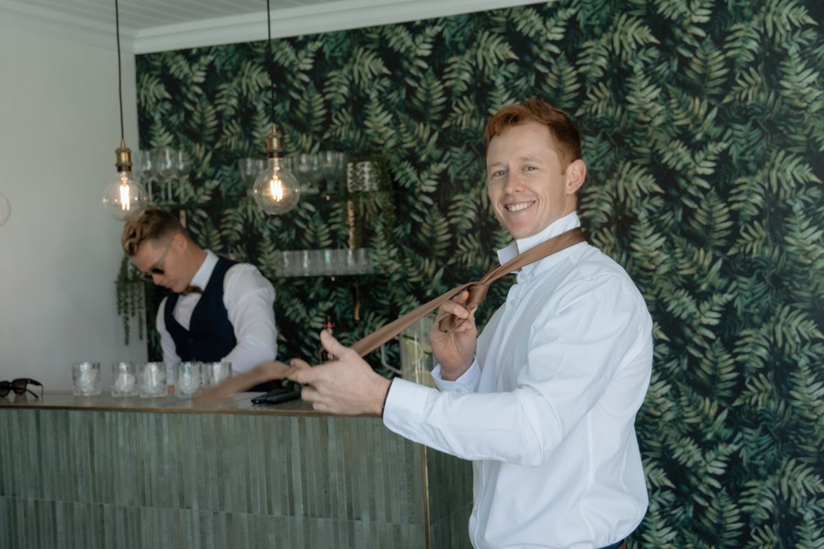 Smiling redhead bartender in white shirt adjusts tie while colleague works at a stylish bar with tropical wallpaper.