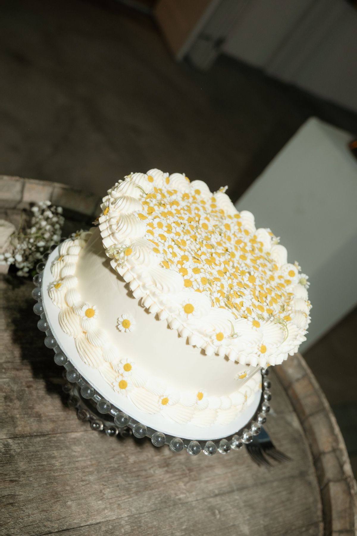 Two-tier white wedding cake decorated with yellow daisy flowers displayed on a rustic wooden barrel.