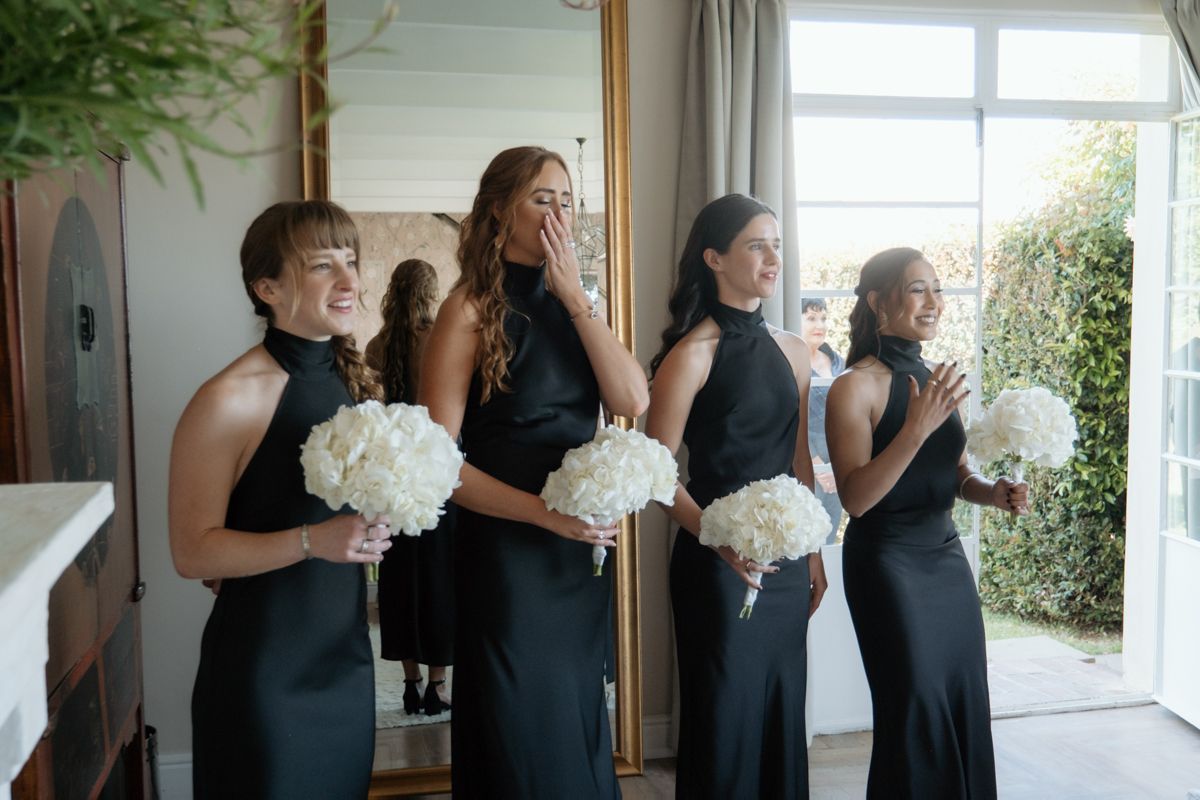 Four bridesmaids in black halter dresses react with surprise and joy while holding white hydrangea bouquets.