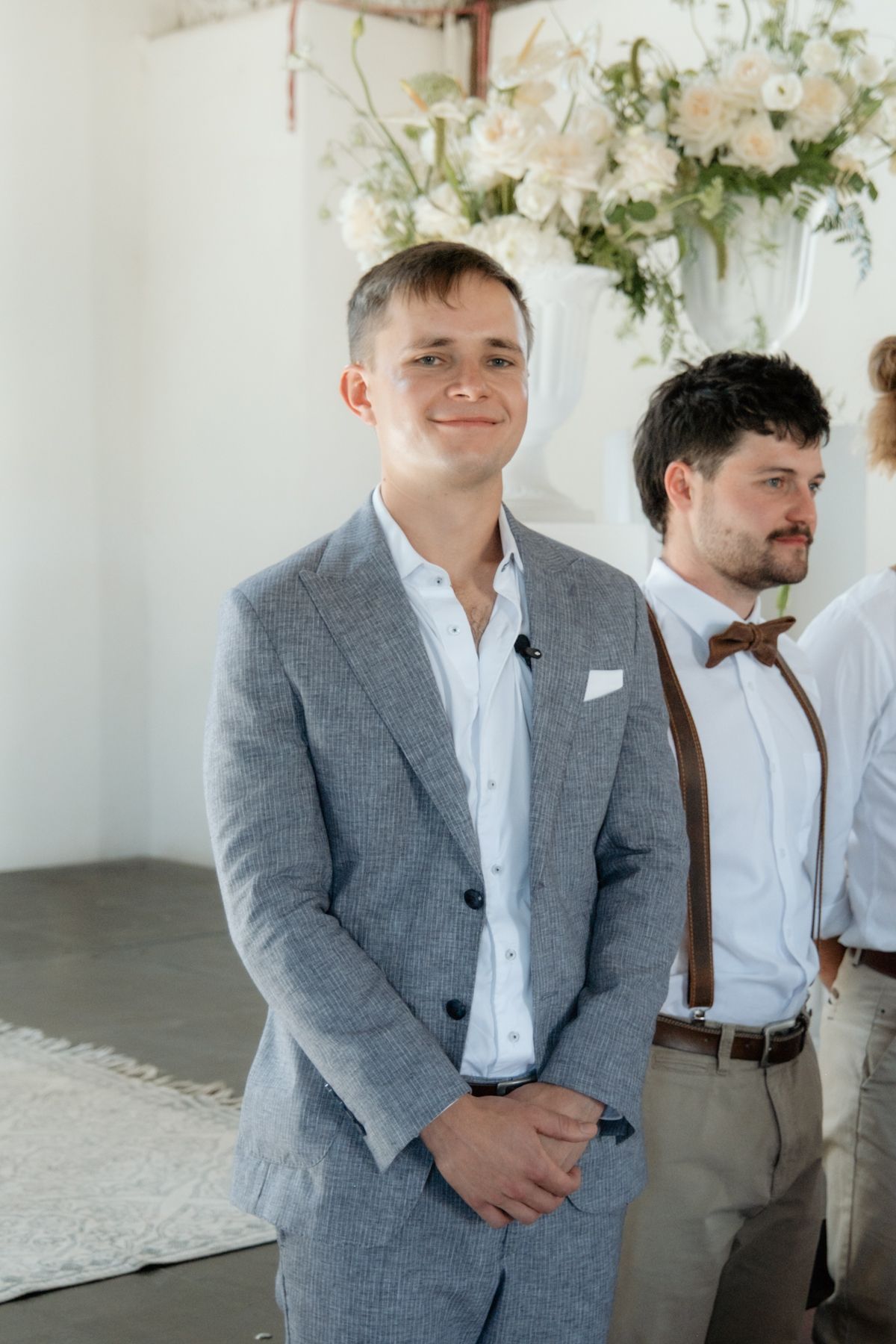 Smiling groom in grey suit stands at altar with groomsman in suspenders at floral wedding ceremony.