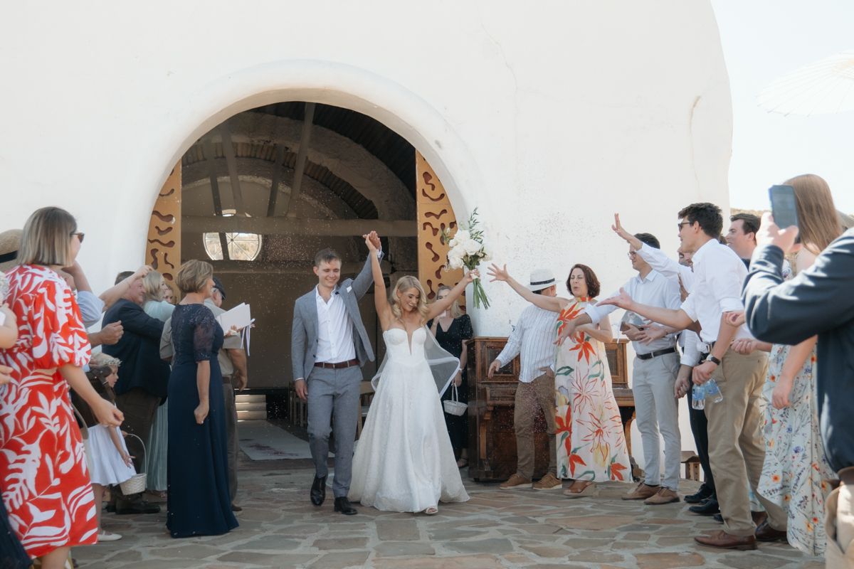 Bride raises arms in joy as she exits a Mediterranean chapel with groom, surrounded by cheering wedding guests.
