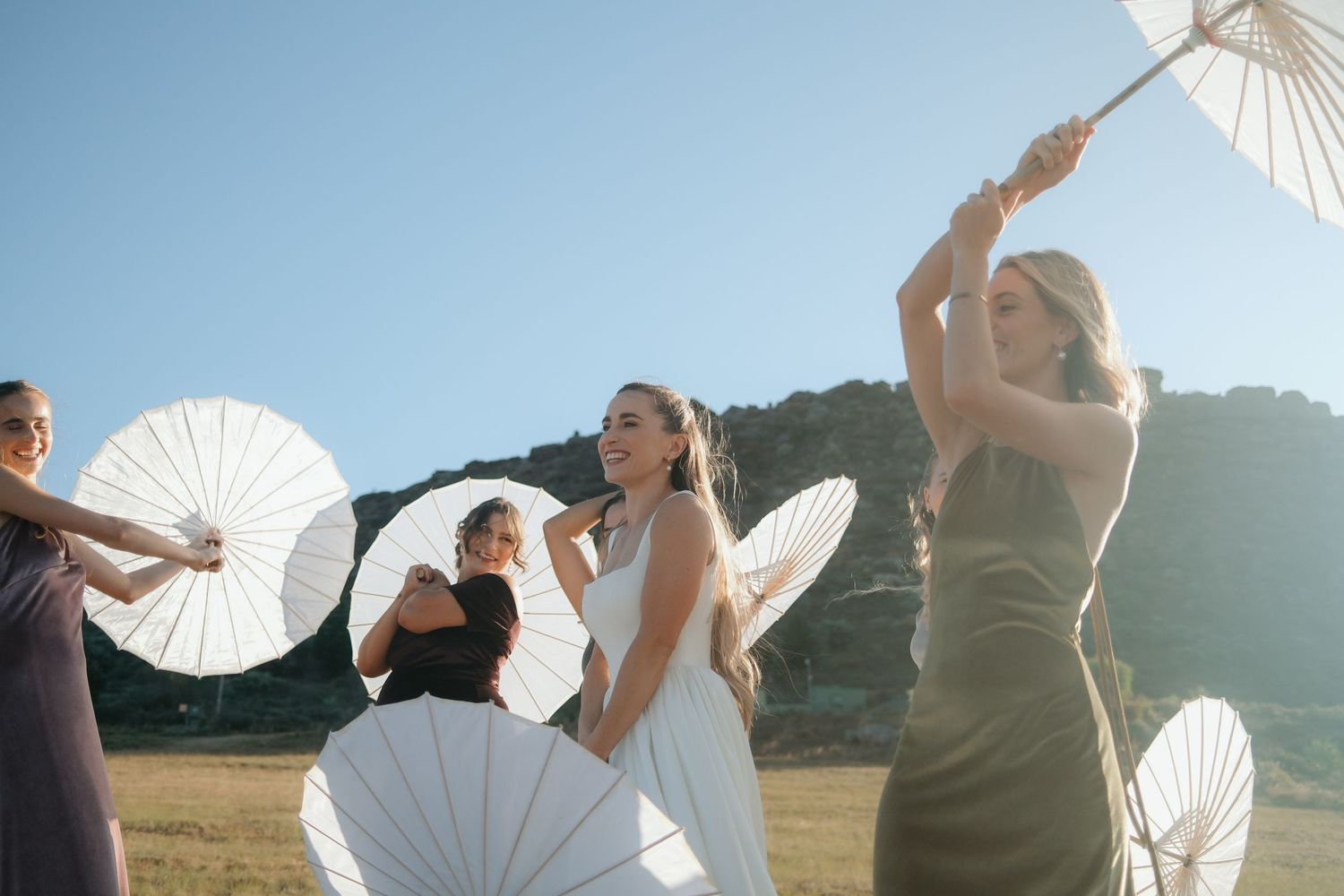 Bridesmaids in elegant dresses twirl white parasols outdoors under a clear blue sky with scenic hills in the background.