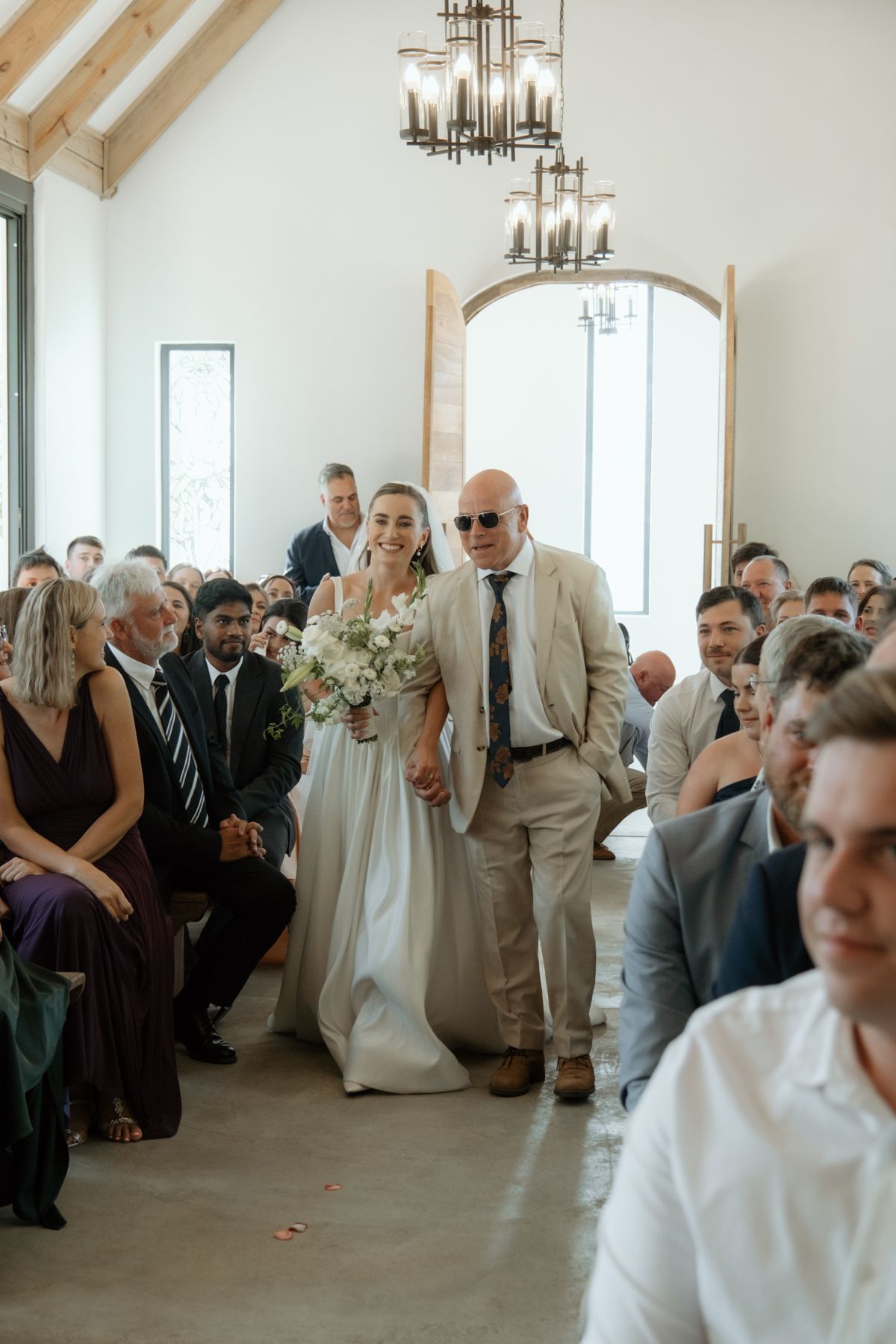 Bride walks down the aisle with her father in a bright white wedding chapel.