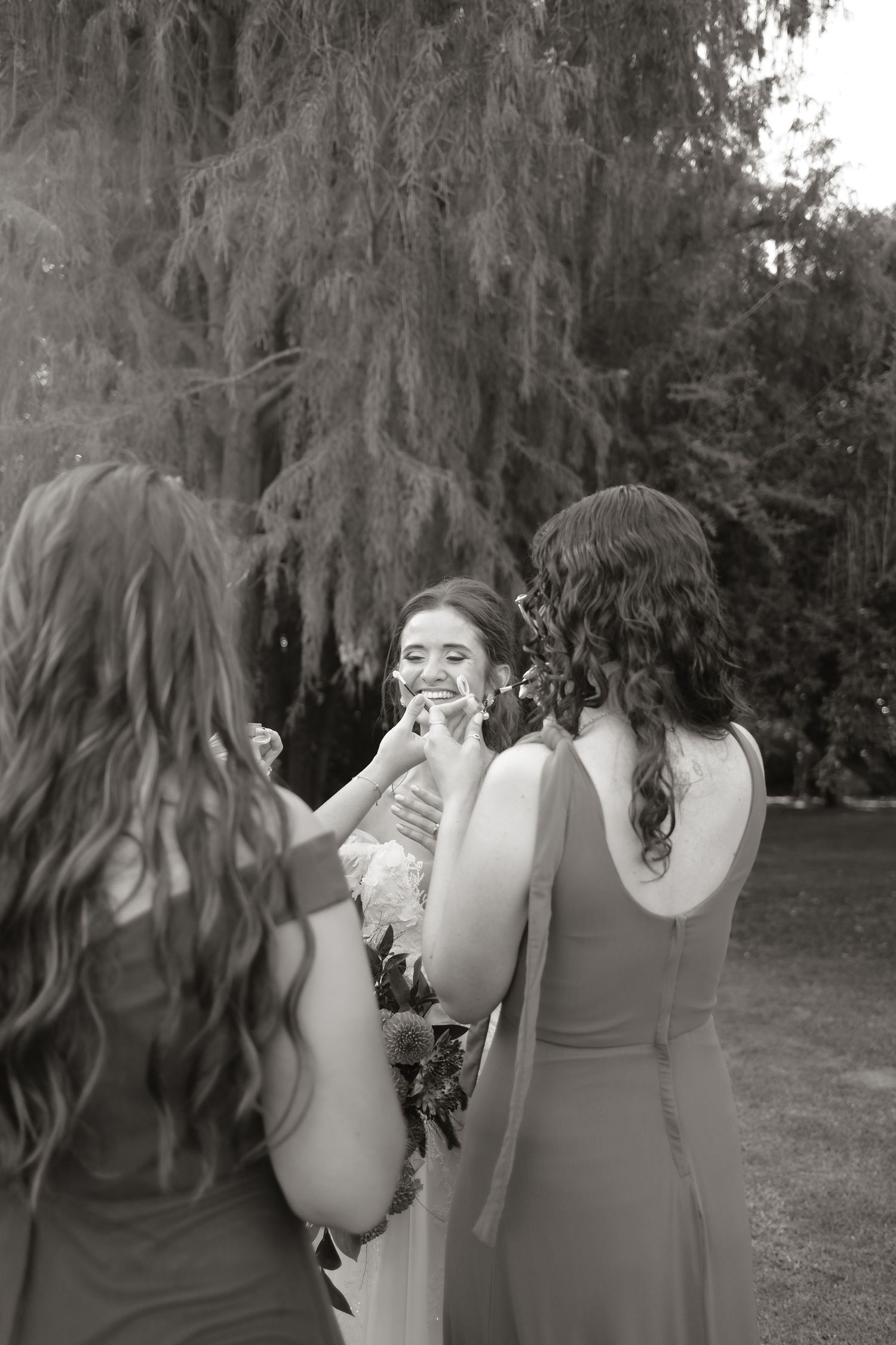 Black and white photo of bridesmaids tenderly touching smiling bride's face in a garden setting.