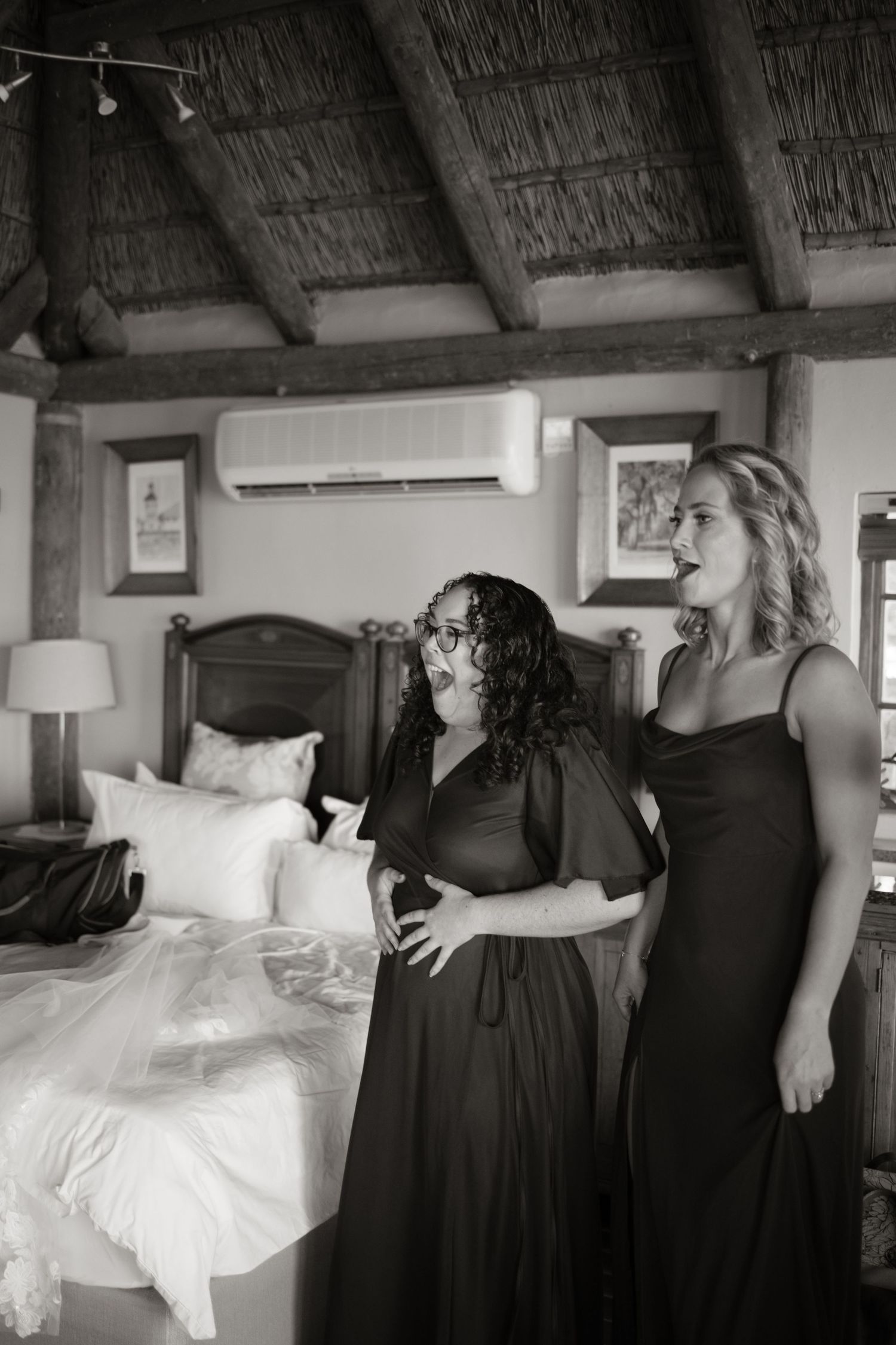 Two bridesmaids in black dresses laugh together in rustic thatched-roof bedroom before wedding ceremony.