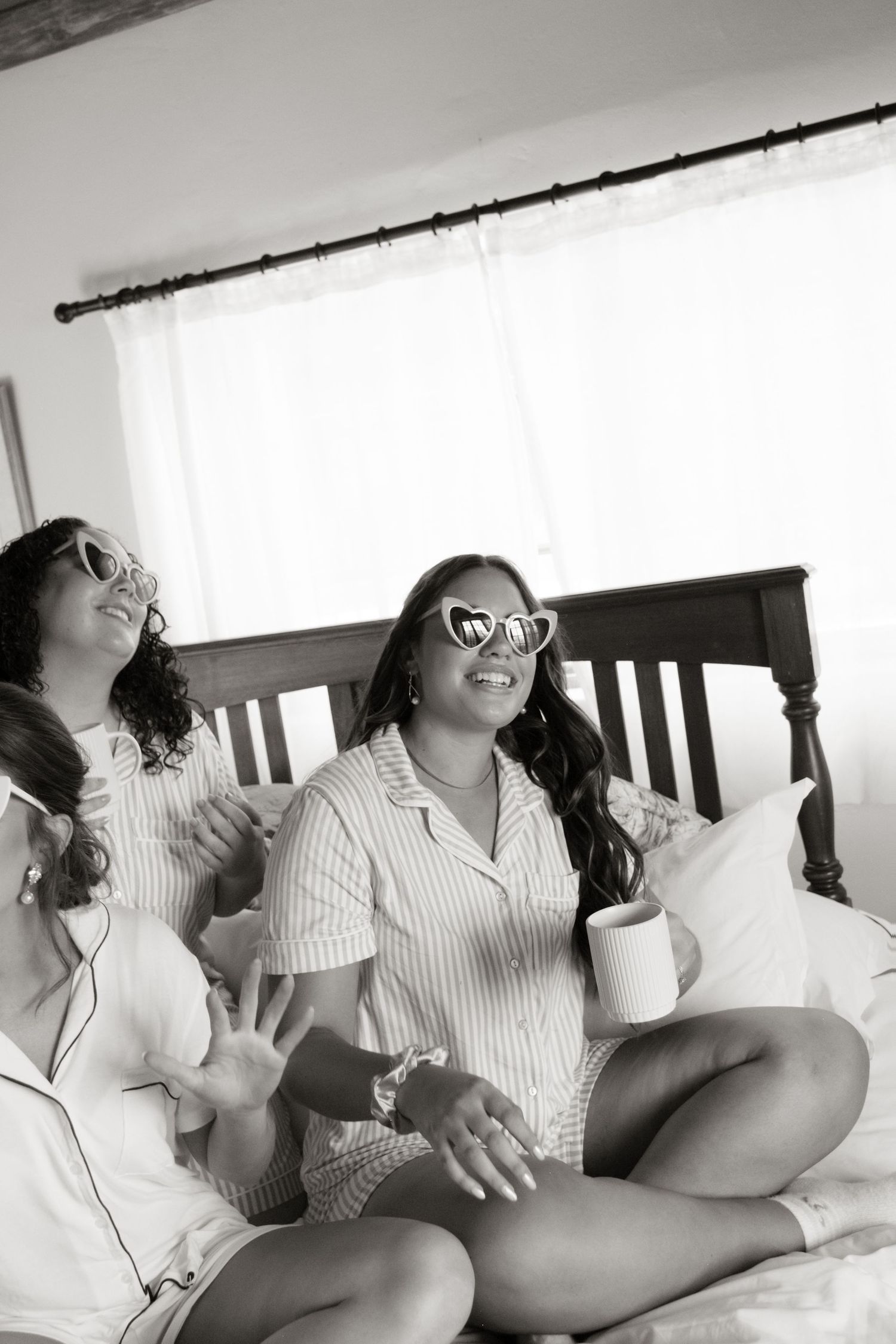 Bridesmaids in striped pajamas relax on a bed with heart sunglasses and coffee mugs before the wedding.