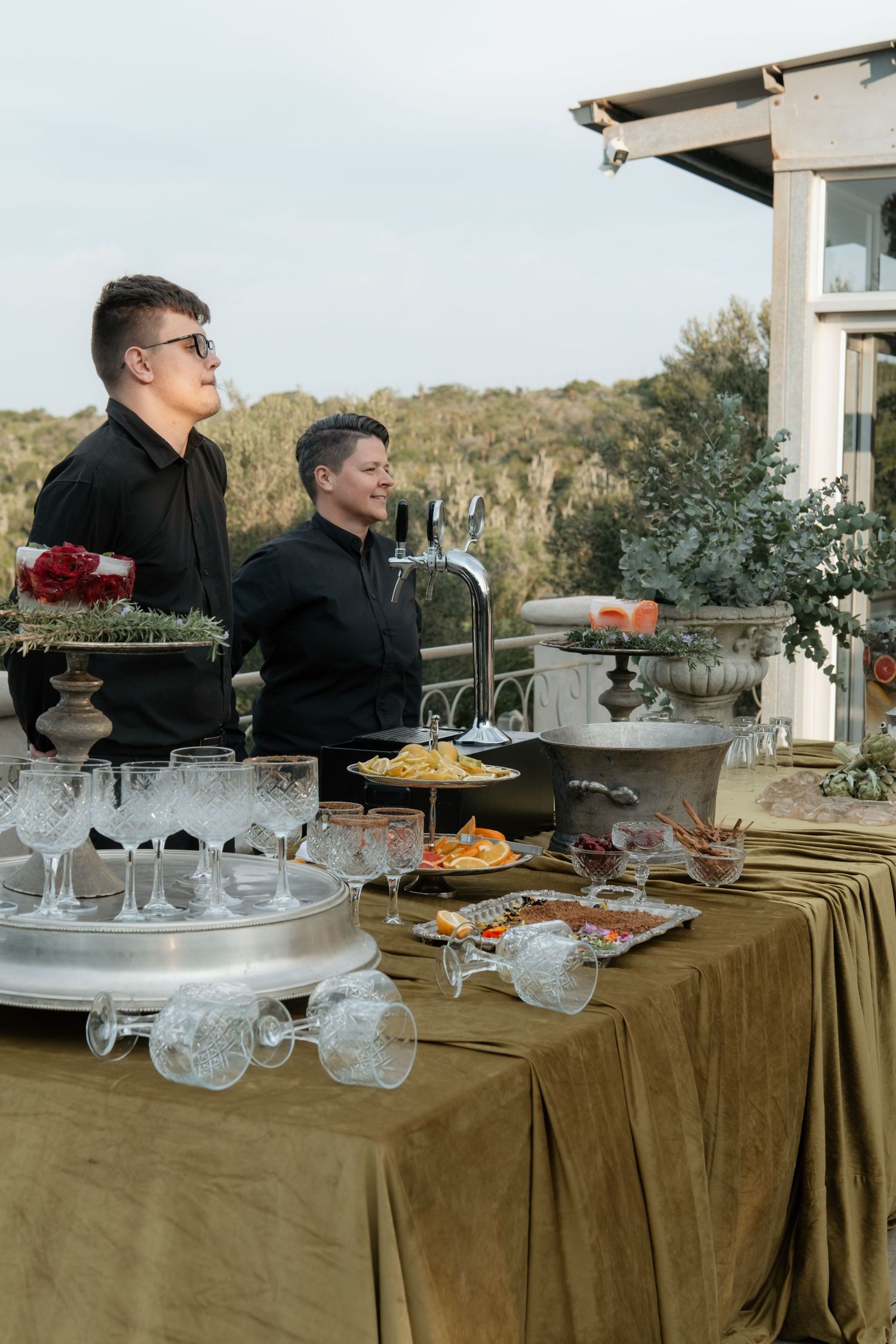 Two servers in black stand behind elegant outdoor bar table with crystal glasses and appetizers.