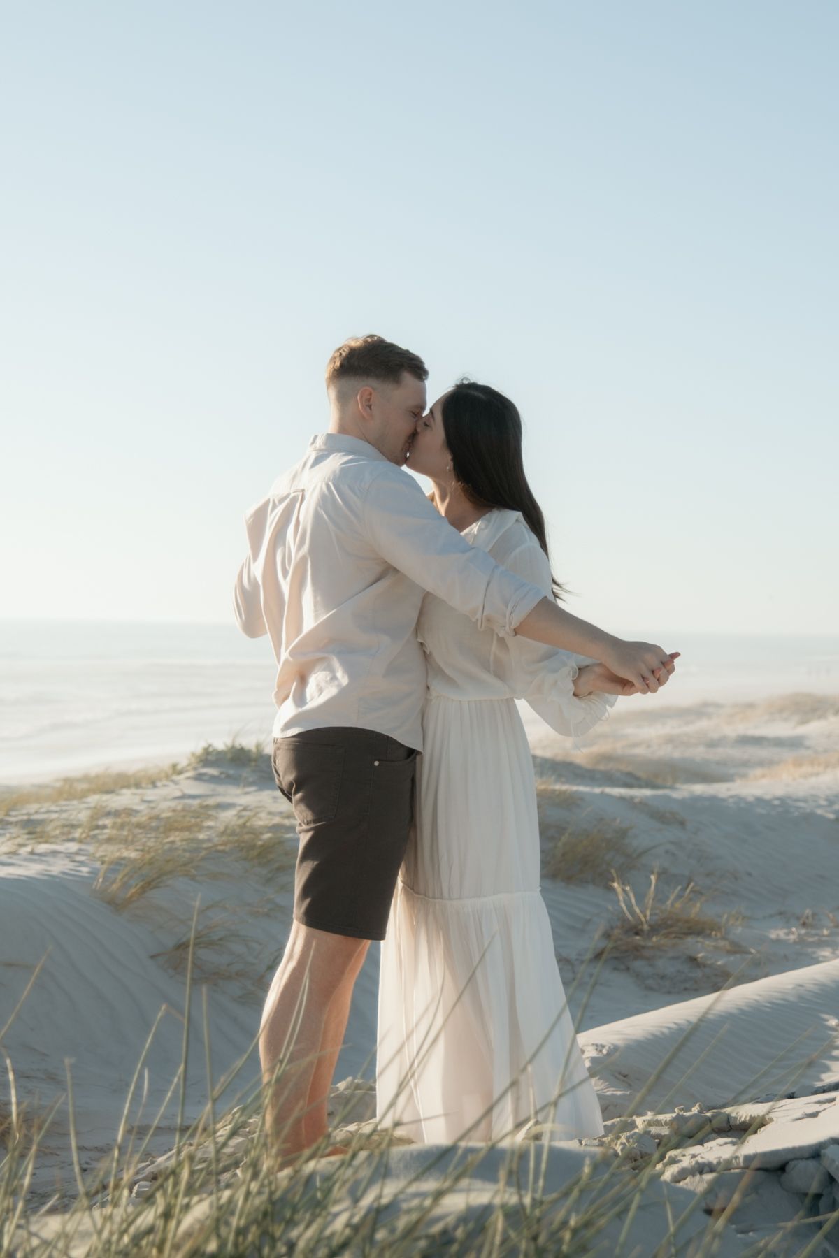 A romantic couple embraces while standing on sandy dunes during golden hour at the beach.