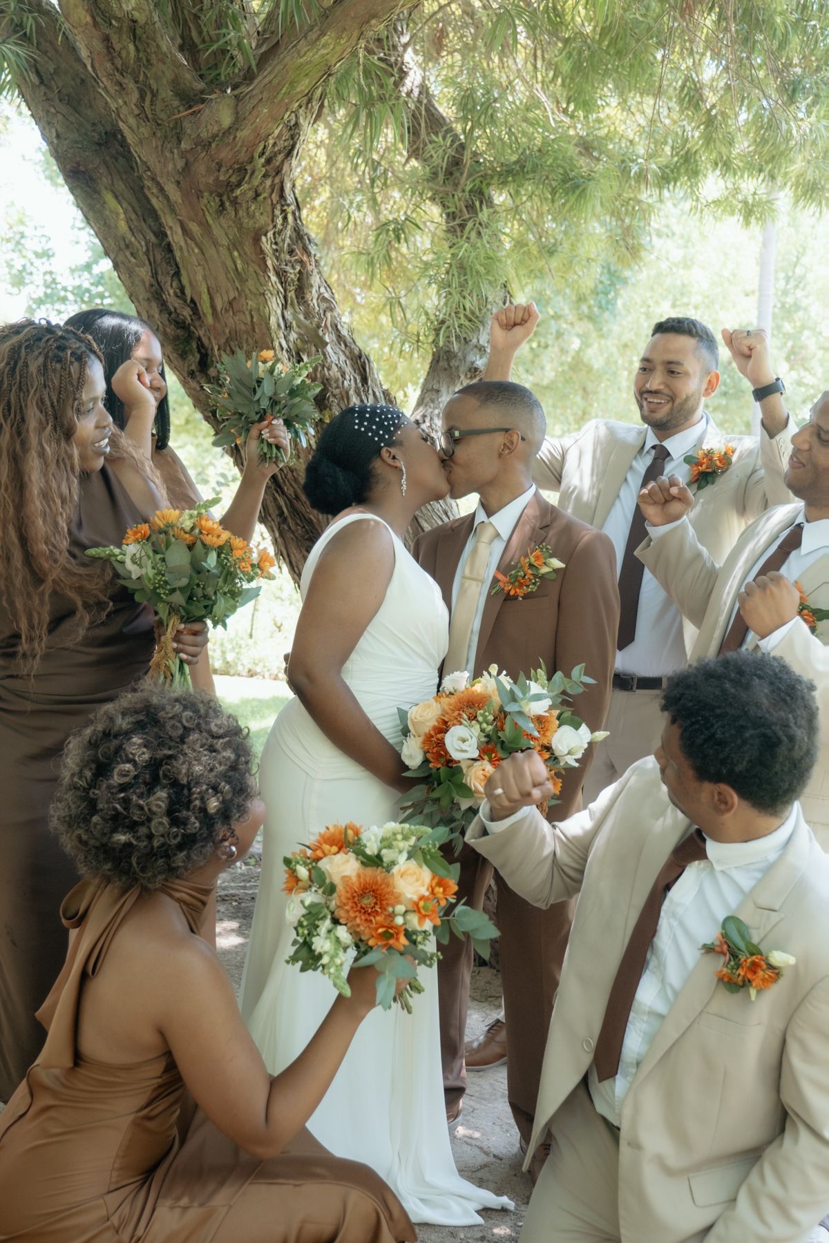 Bride and groom share a kiss surrounded by cheering wedding party in brown and tan attire under a shady tree.