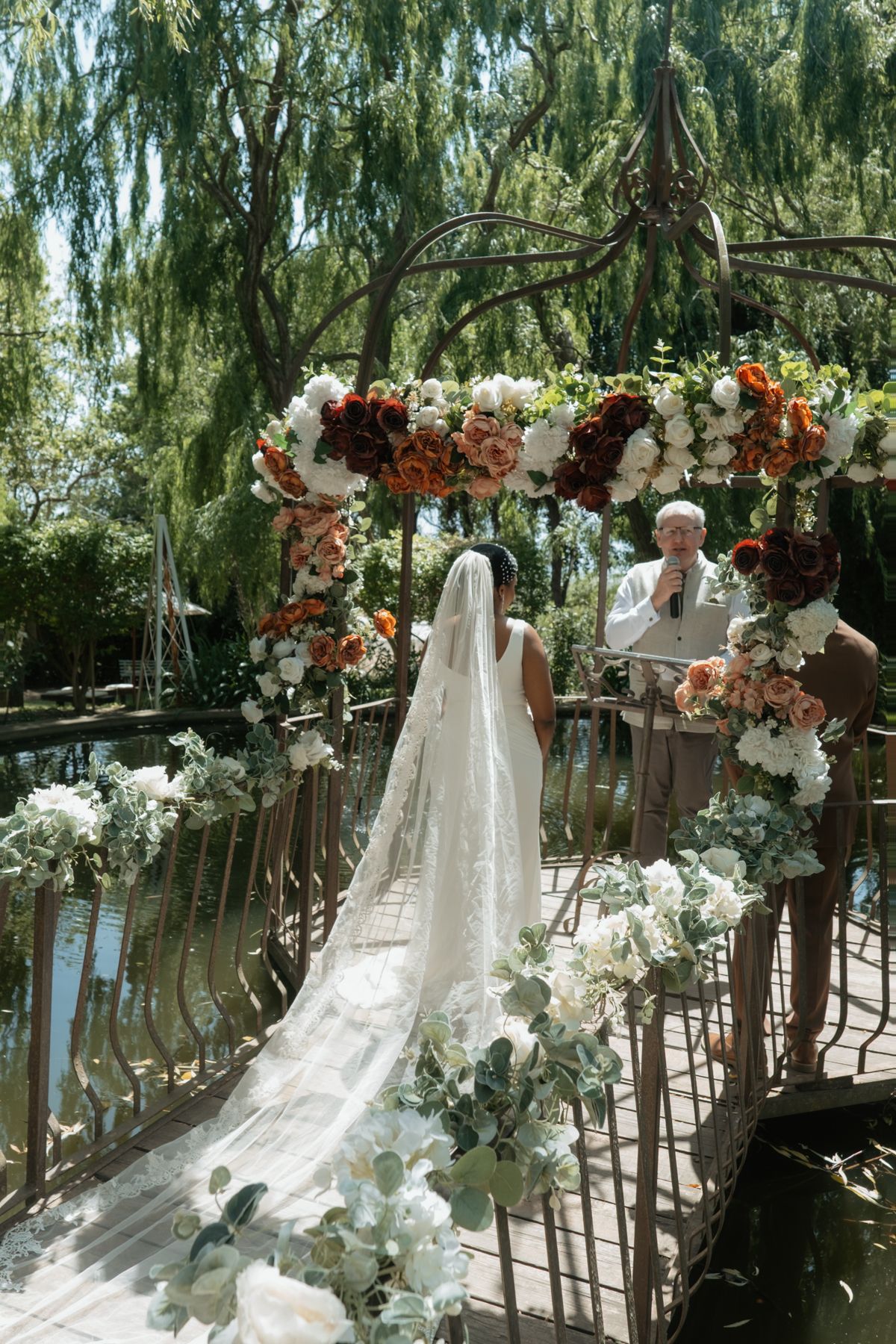 Bride in lace veil stands before flower-adorned arch gazebo at outdoor wedding.