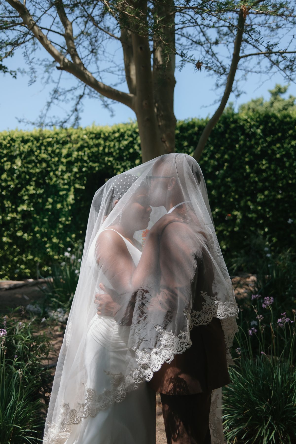 Romantic wedding photo of bride and groom kissing hidden beneath a flowing lace bridal veil outdoors.