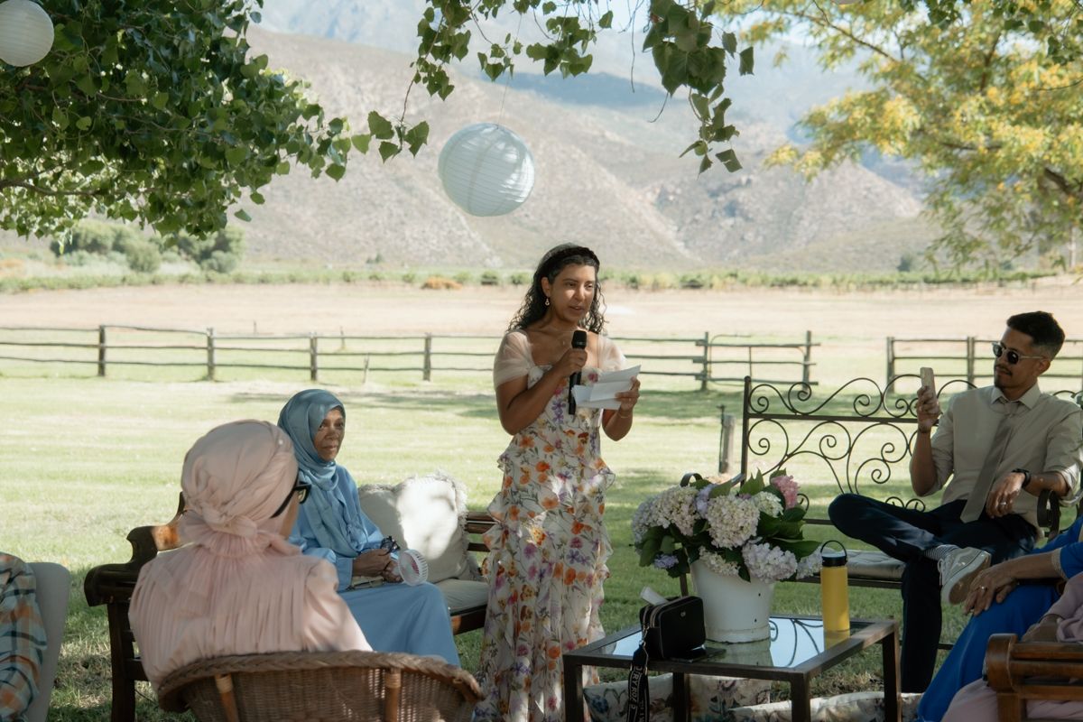 Woman in floral dress speaks into microphone at outdoor wedding reception with mountain views.