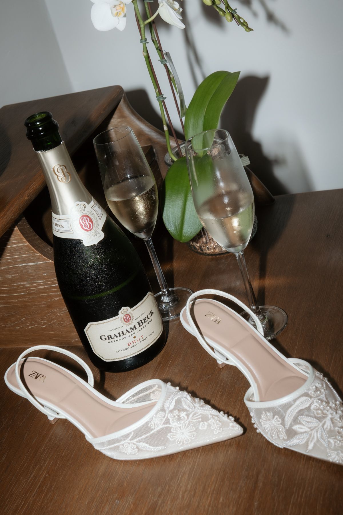 White lace bridal heels displayed beside Graham Beck champagne bottle and filled flutes on wooden table.