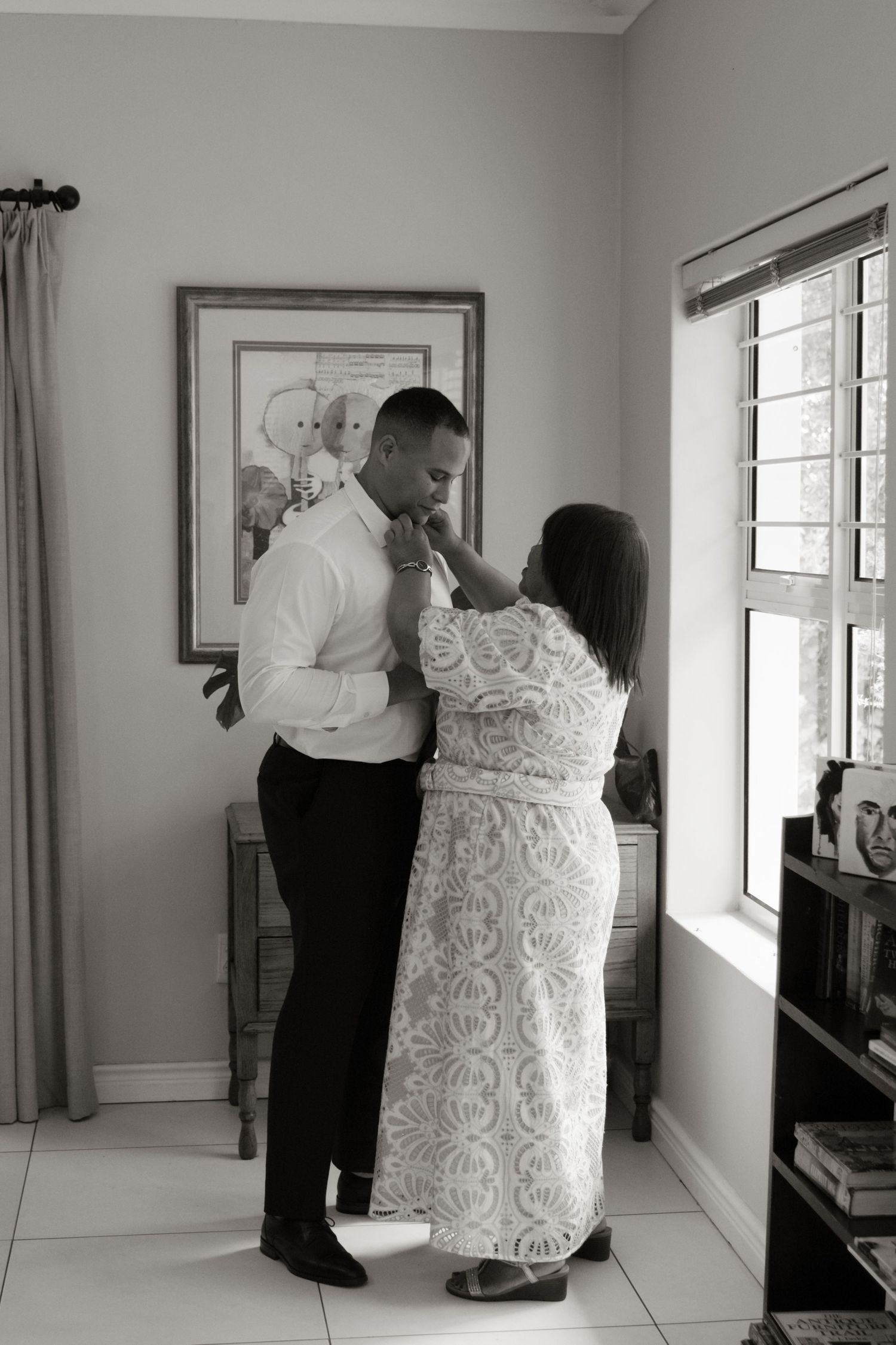 Black and white photo of a couple sharing a tender moment, woman in lace dress touching mans face indoors.