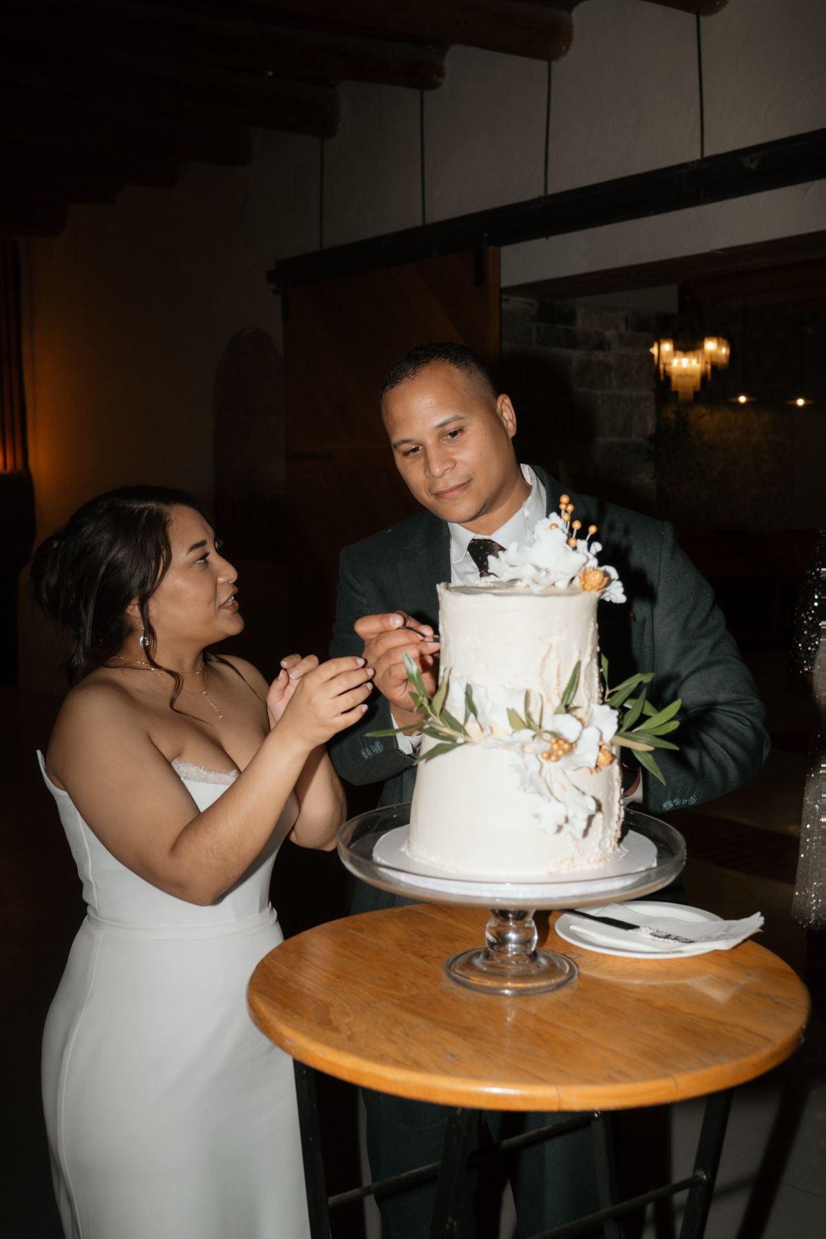 Bride and groom cut their elegant two-tier white wedding cake decorated with flowers and greenery.