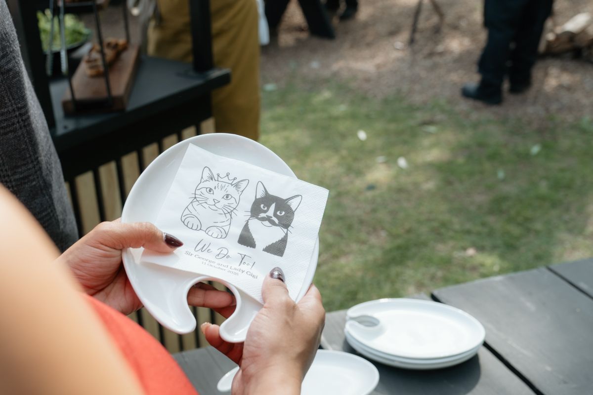 Hands holding a decorative white cat-shaped plate with illustrated cats at an outdoor wedding event.