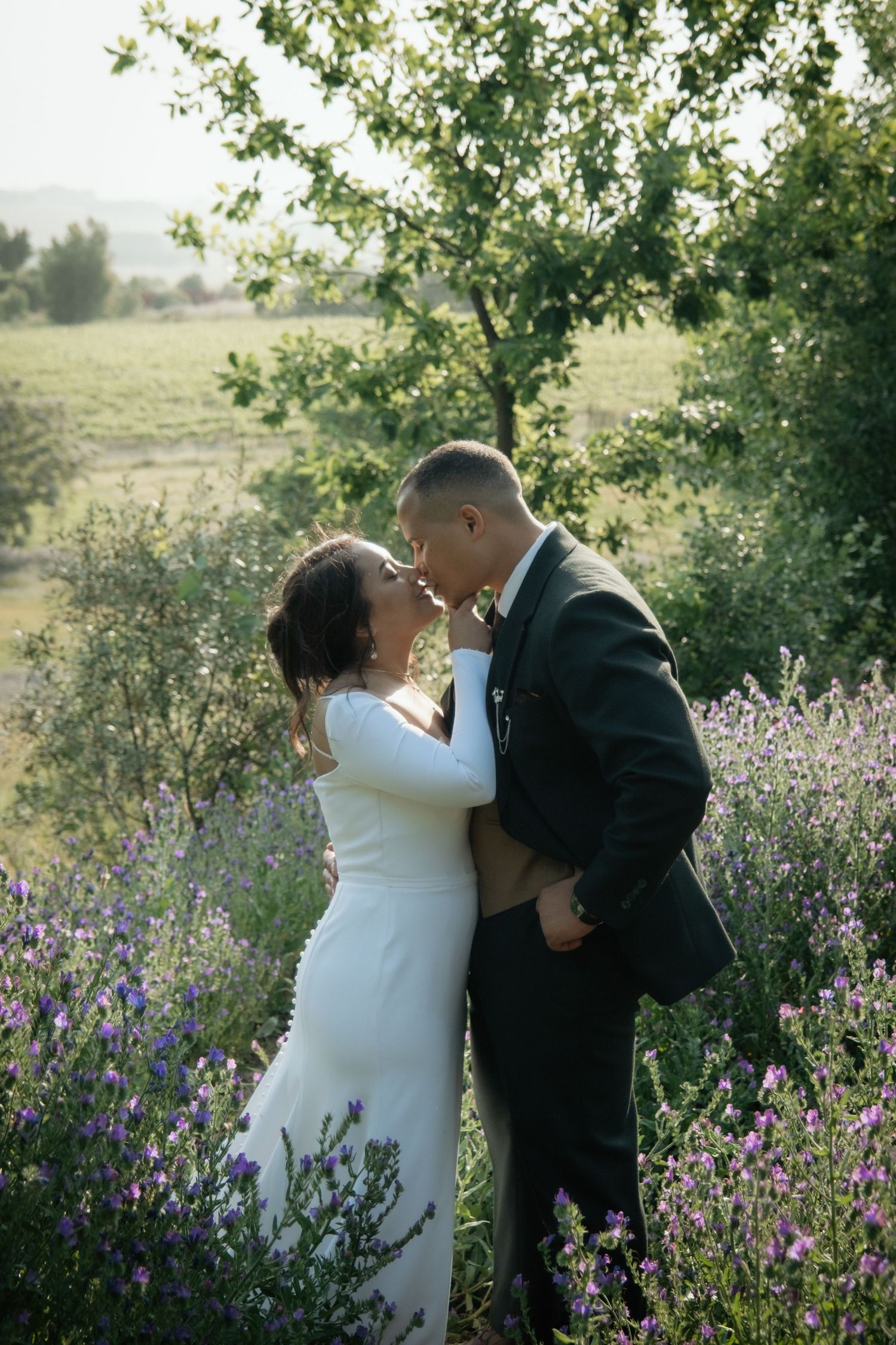 Bride and groom sharing a romantic kiss surrounded by lavender flowers in a scenic outdoor setting.