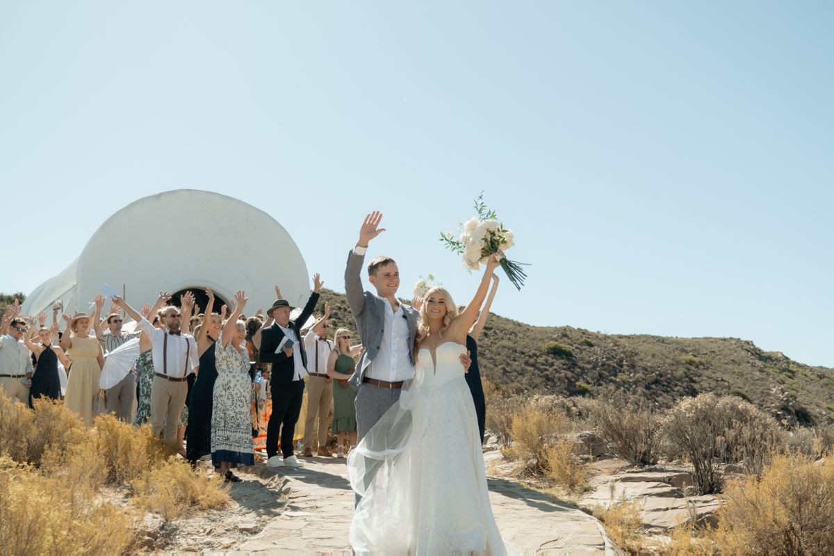 Bride and groom raise arms in joy after wedding ceremony with guests cheering in scenic outdoor setting.