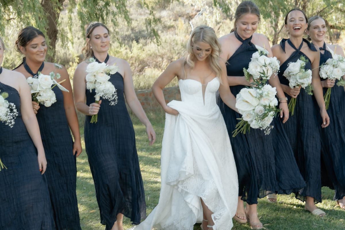 Bride in strapless white gown walks with bridesmaids in navy dresses holding white floral bouquets outdoors.