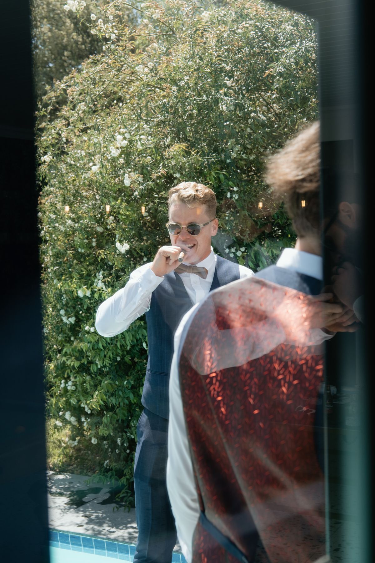 Dapper groom in navy vest and sunglasses adjusts his bow tie outdoors beside lush green hedges.