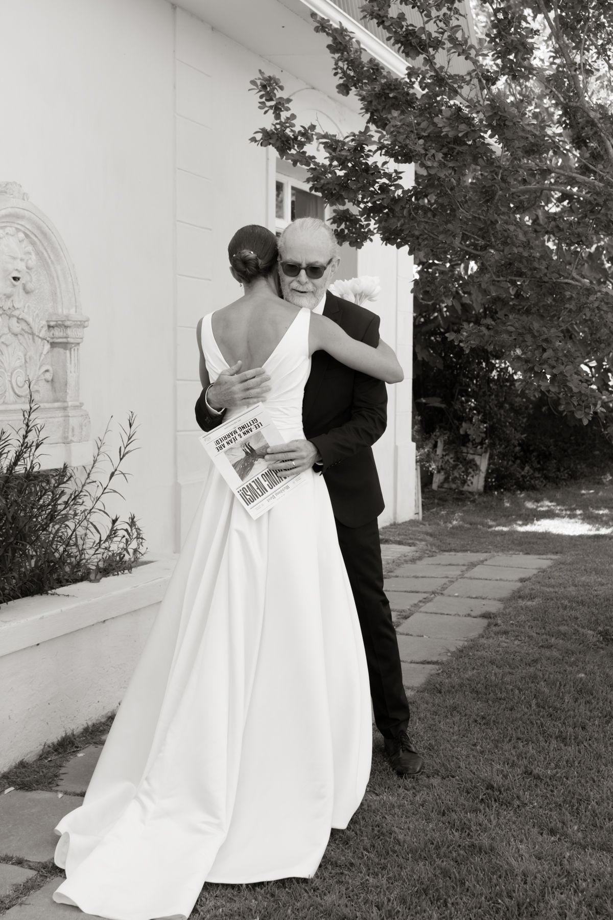 Black and white photo of bride in backless gown sharing tender embrace with father outdoors.