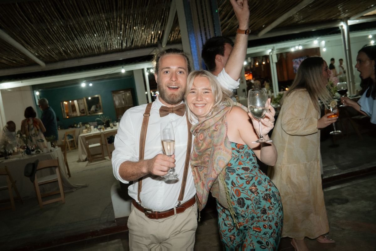 Smiling couple in wedding attire holding champagne glasses and celebrating at an indoor wedding reception party.